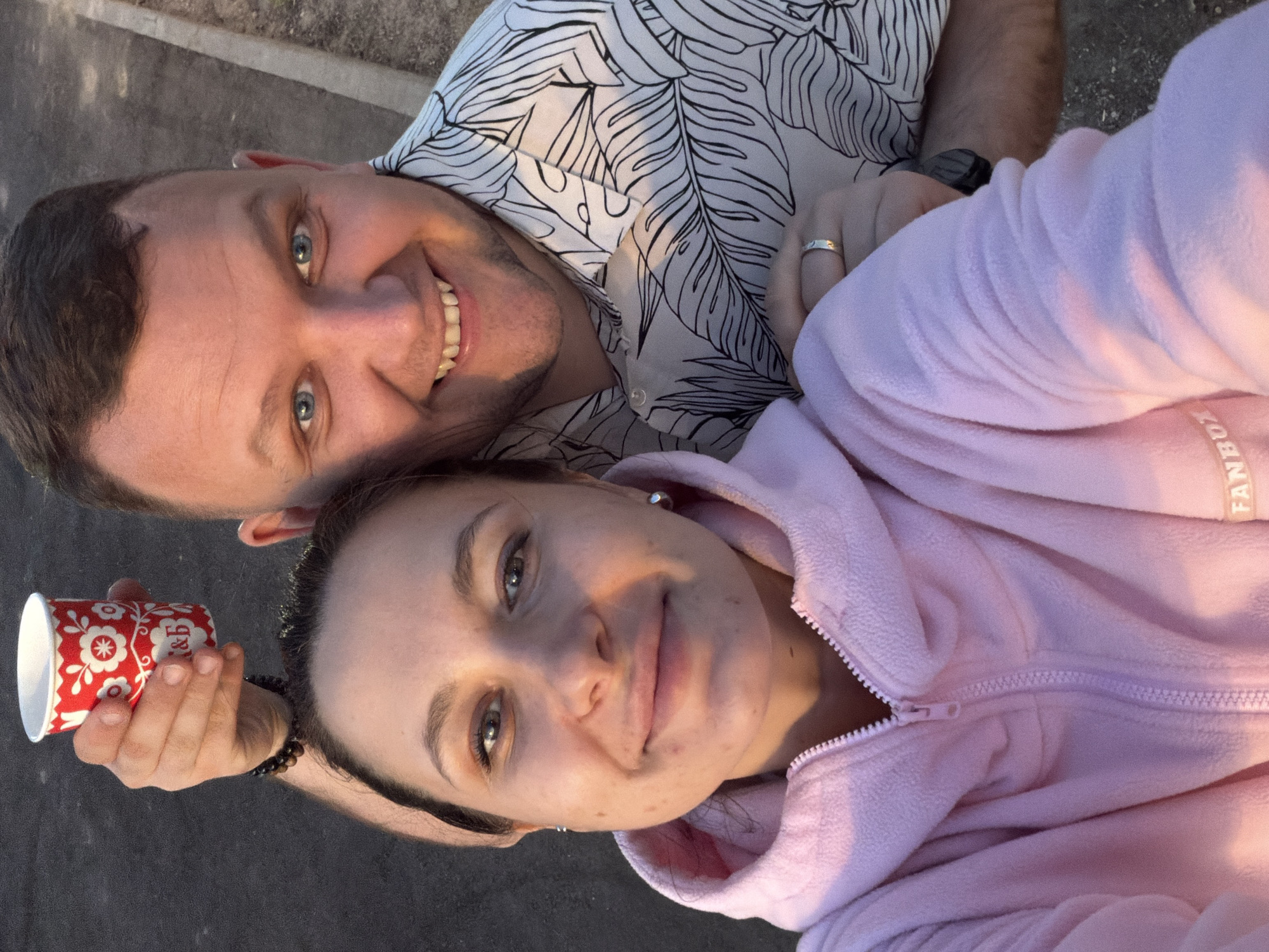 This image captures a close-up outdoor selfie of two individuals, one female and one male, in warm natural light. The female, in the foreground on the left, appears to be in her late 20s to early 30s, with dark hair and a gentle smile directed at the camera. She is wearing a pink fleece hoodie with a zipper, and the text "FANBOX" is visible on her left sleeve. She has small silver stud earrings. The male, positioned behind and to the right of the female, appears to be in his late 20s to 30s, featuring short dark hair, blue eyes, and a broad smile. He is wearing a light-colored button-down shirt with a black tropical leaf pattern. His right arm is around the female, and his left hand is raised above her head, holding a small red and white paper cup adorned with a floral design and the Cyrillic letters "К&Б." A dark beaded bracelet is on his left wrist, and a silver band ring is on his left ring finger. The background is blurred, showing a dark asphalt surface and a light-colored curb or pathway edge. The lighting suggests either early morning or late afternoon. The "К&Б" marking on the cup, widely recognized as belonging to the "Krasnoe & Beloe" retail chain, indicates the location is in Russia, consistent with the prompt's specification of Moscow.