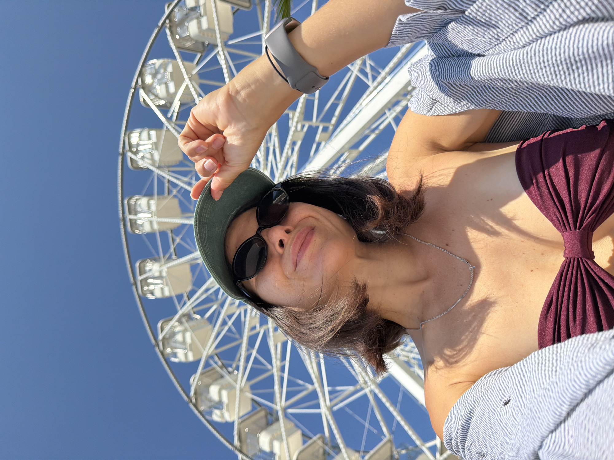 A low-angle, full-body outdoor selfie shows a woman with shoulder-length brown hair, wearing a dark red bikini top, a grey and white striped button-up shirt worn open, sunglasses, and a green baseball cap. She is smiling with her head tilted back and one hand resting on the brim of her cap. A silver necklace is visible around her neck, and a grey fitness tracker is on her left wrist. Behind her, a large white Ferris wheel with several white gondolas is silhouetted against a clear blue sky. The overall lighting suggests a sunny day.
