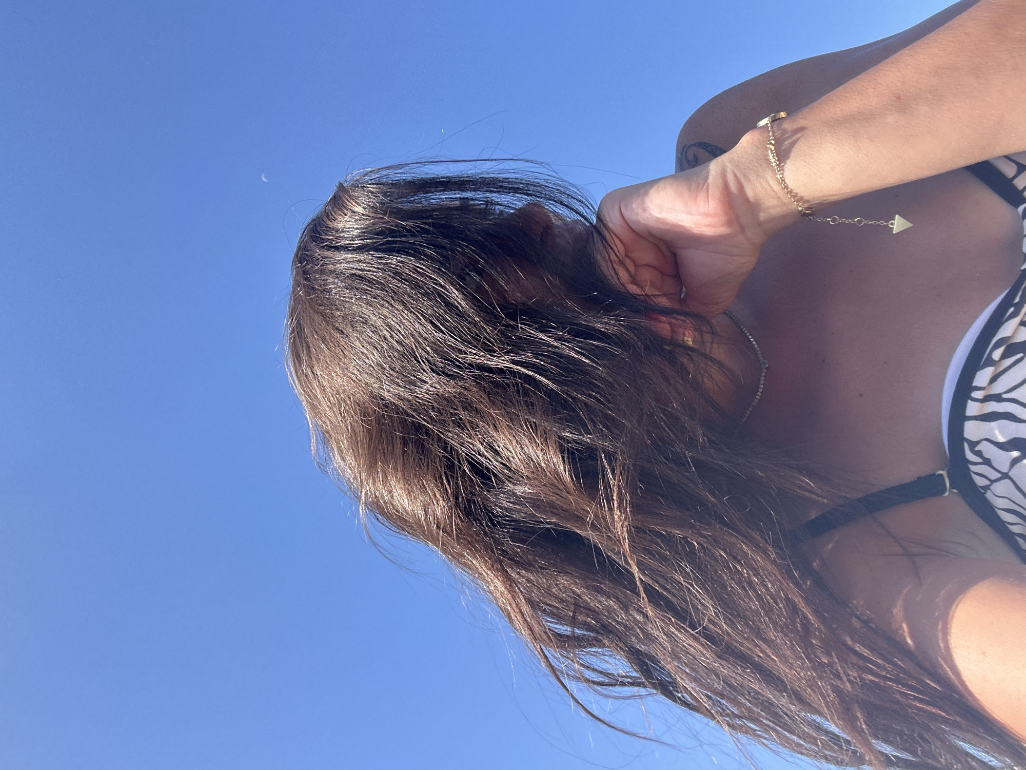 A low-angle medium shot captures a person from the chest up, looking to the right with their head tilted upwards against a clear, bright blue sky. Their long, dark brown hair is windblown, obscuring part of their face. They are wearing a black and white patterned bikini top and a delicate gold necklace. A gold bracelet with a triangular charm is visible on their right wrist. The person's right hand rests against their cheek, with a tattoo visible on their right shoulder. The scene appears to be outdoors, with the bright sky suggesting a sunny day. The location is Cala Lliteres, Spain.