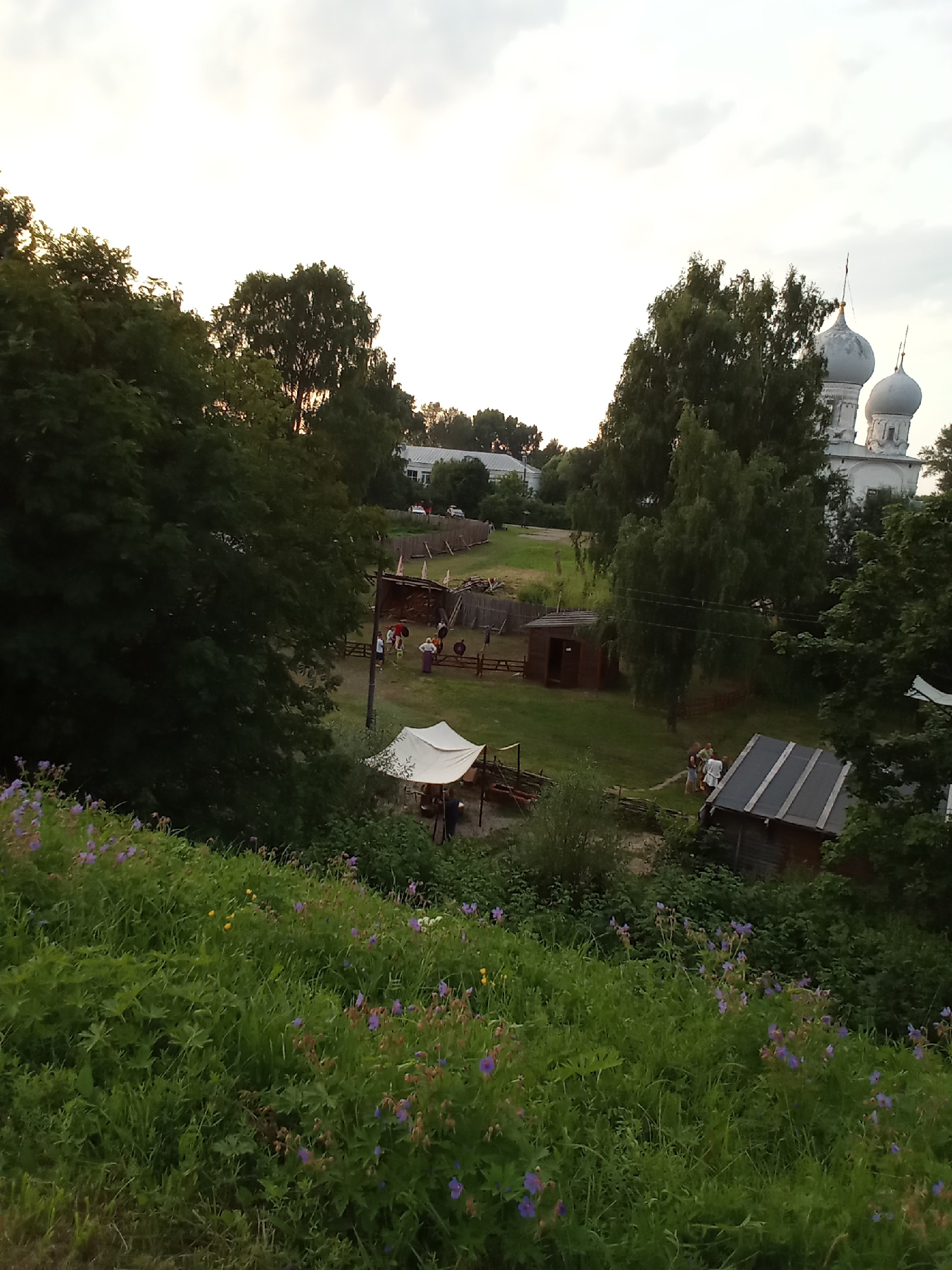 The image captures an outdoor scene in what appears to be an open historical reenactment or village area, likely in Belozersk, Russia. The foreground is dominated by a dense, green grassy slope dotted with small purple and yellow wildflowers. In the middle ground, a cluster of rustic wooden structures and a white tent are visible, set against a backdrop of lush green trees. Several people, dressed in what might be period clothing, are scattered around this area, with some appearing to be interacting near the tent and other structures. A fenced enclosure, possibly for animals or a pathway, is also present. In the background, the distinctive onion domes of a white church rise above the trees, indicating a significant architectural landmark. The sky is overcast with a bright, diffused light, suggesting it is either early morning or late evening. The overall impression is of a tranquil, historical setting within a natural landscape.