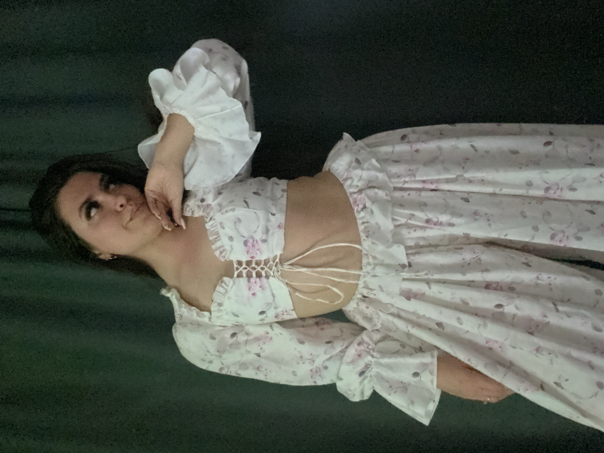 A woman with dark hair is lying on a dark green surface. She is wearing a floral print dress with ruffled sleeves and lace-up detailing around the midriff. Her left hand is resting on her neck, and her right hand is resting on the floor near her waist. She is looking upwards and to the left of the frame. The location in Chaikovsky, Russia, is not determinable from the image.