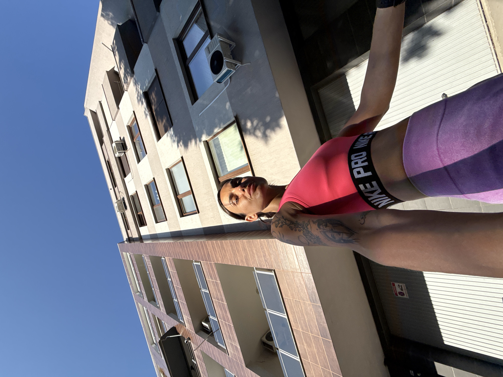 An outdoor, low-angle, full-shot photograph captures a woman leaning against a building in Pleven, Bulgaria. The woman, positioned on the right side of the frame, wears a pink sports bra with "NIKE PRO" branding and purple leggings. Her left arm, adorned with tattoos, rests against her body, while her right arm extends forward, partially out of frame. The background features a multi-story building with several balconies and windows. An air conditioning unit is visible on one of the building's exterior walls. The sky above is a clear, bright blue. Sunlight casts shadows across the scene, indicating it is daytime.