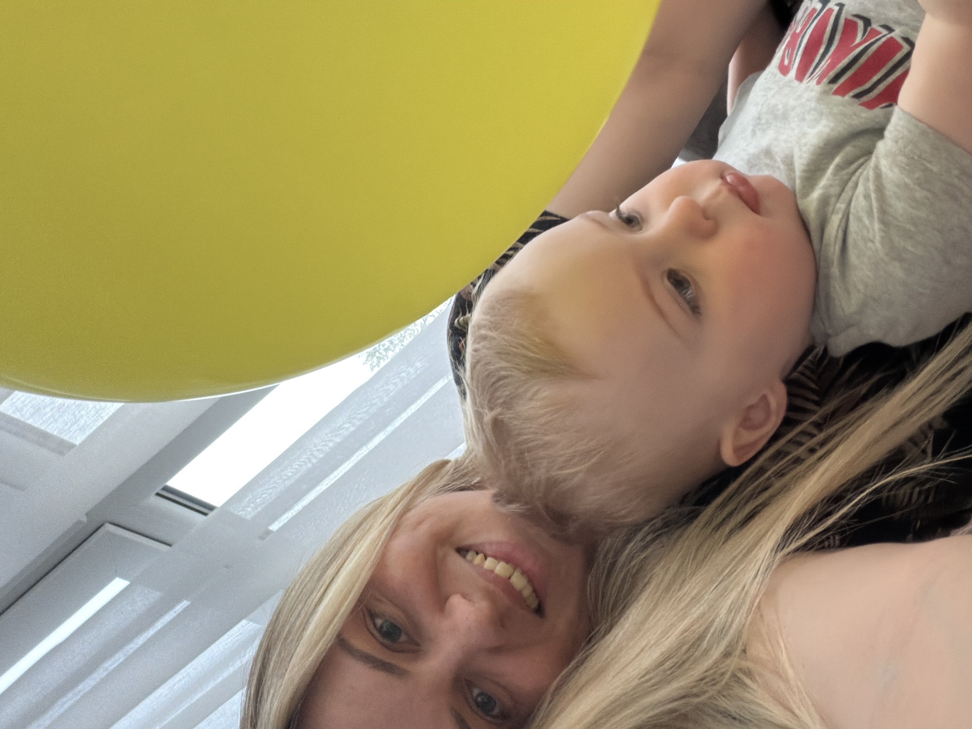 A tilted, close-up, indoor photograph depicts a Caucasian woman with blonde hair and a Caucasian child. The woman is smiling and the child is looking to the upper right. A large yellow balloon is partially visible in the upper left corner. The background shows a window with white blinds. The location is likely indoors. Information about the specific city Votkinsk, Russia is not discernible from the image.