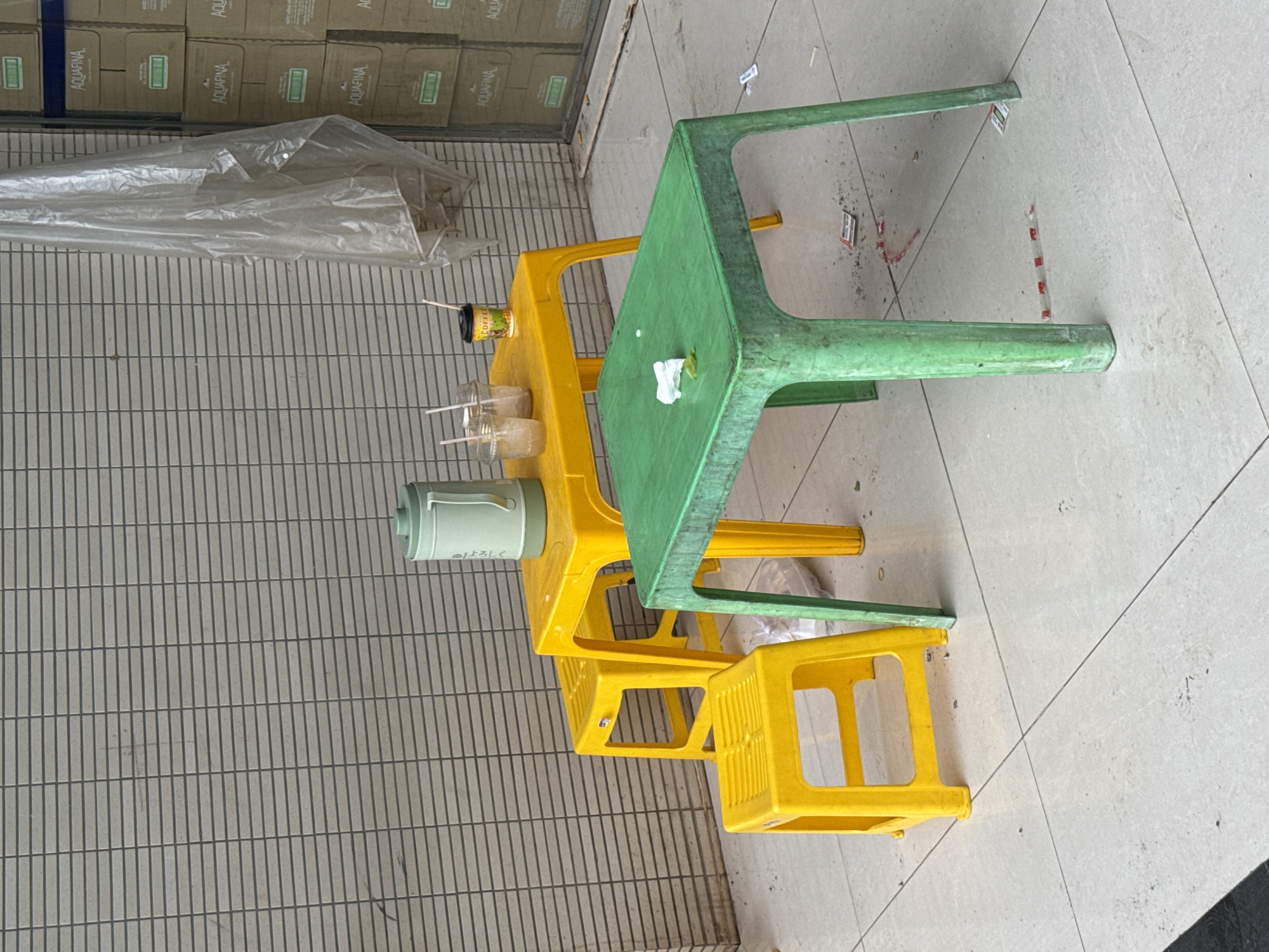 An outdoor scene in Lao Bảo Commune, Vietnam, features two plastic tables and three plastic stools on a tiled floor. A yellow table, elevated by two yellow stools, holds a green thermos, three clear plastic cups with straws, and a small coffee cup. A green table, lower than the yellow one, is positioned in front of it. The environment includes a tiled wall in the background and stacked boxes to the right, with a clear plastic sheet hanging. The area appears to be an outdoor seating area for a small shop or eatery, possibly a roadside establishment. The scene is devoid of any people.