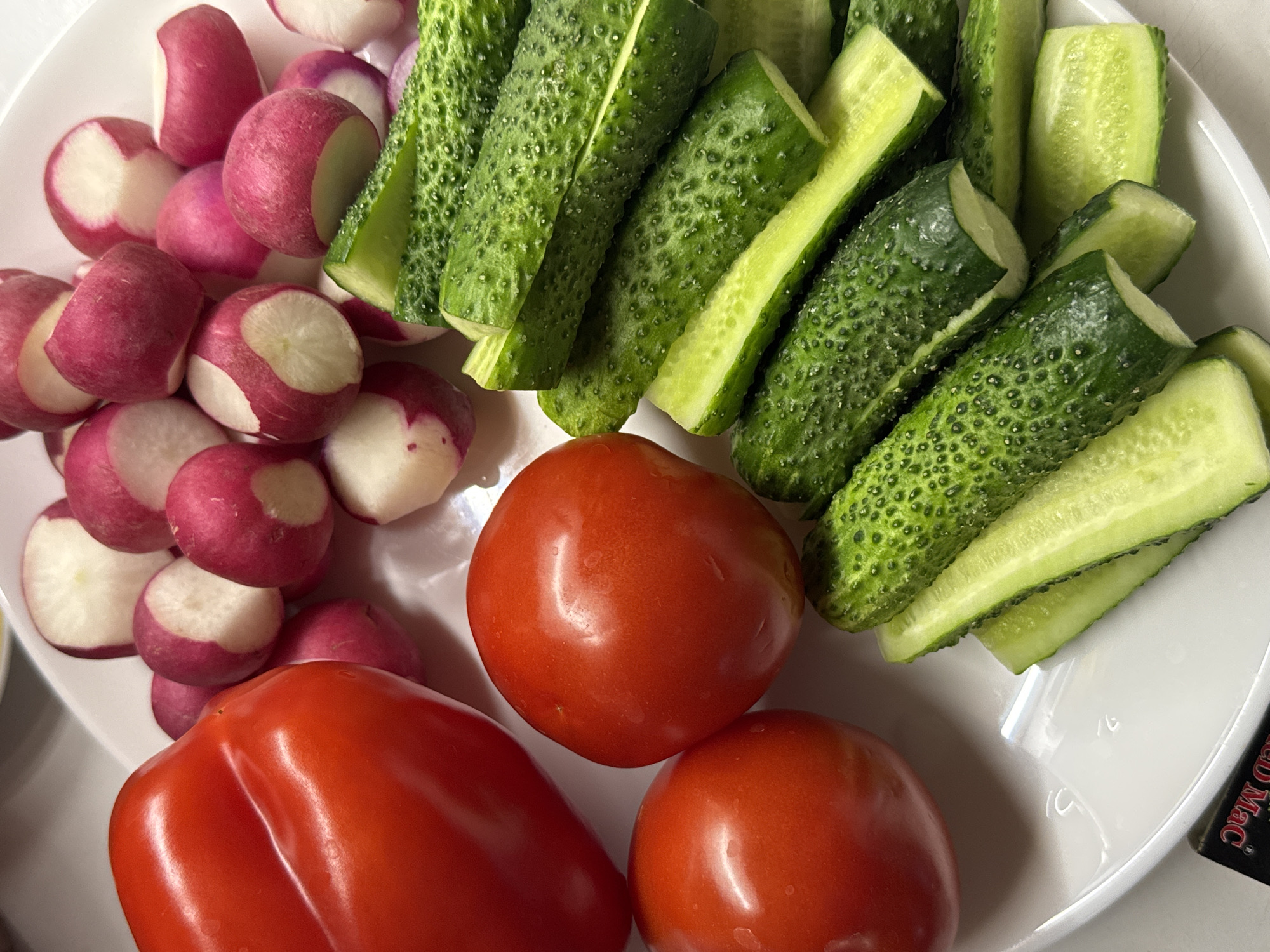 A white plate holds a variety of fresh vegetables. A large, red bell pepper is positioned on the left side of the plate, with two round, red tomatoes to its right. In the upper portion of the plate, a pile of sliced radishes, showing their white insides and pink skin, is visible. The bottom and right portions of the plate are filled with numerous chopped cucumbers, cut lengthwise into spears. The environment is clean and well-lit, suggesting a kitchen or dining setting. The specific location in the city of Unknown, Unknown is not discernible from the image.