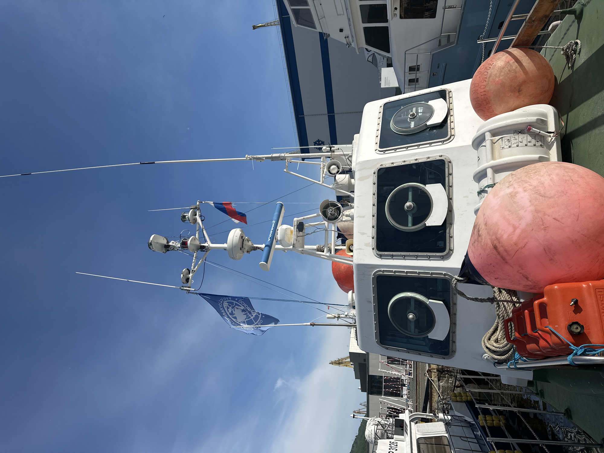 The image shows the deck of a white research vessel docked in the harbor of Petropavlovsk-Kamchatsky, Russia. The vessel has a raised bridge with large windows, several antennas and radar equipment on the roof, and two flags flying: a blue flag with a globe emblem and a Russian flag. There are also two large orange buoys on deck, a red jerrycan, and coiled rope. In the background, other boats and industrial buildings can be seen, along with a clear blue sky with some wispy clouds. The water in the harbor is visible. The overall scene is one of a working port on a sunny day.