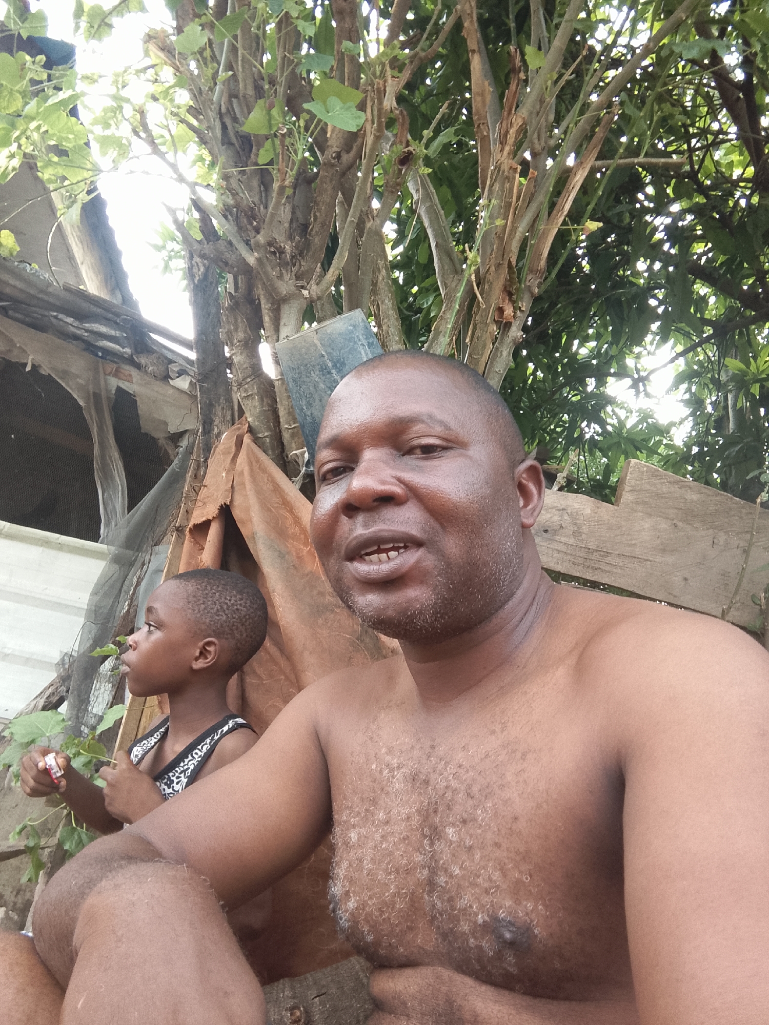A close-up shot shows an adult male and a young child outdoors in what appears to be a less developed area, possibly in the city of Unknown, Unknown. The adult male, who is bald and shirtless, looks directly at the camera. His expression is neutral, with a slight smile. The child, partially visible on the left, wears a patterned tank top and looks off to the side, away from the camera, and is holding some vegetation. Behind them, there are makeshift structures consisting of materials like tarps and wood, along with dense foliage and trees. The lighting suggests it is daytime.