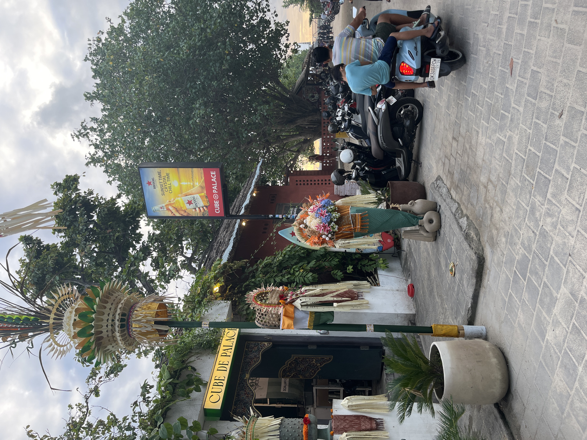 The image shows a street scene in Kuta, Indonesia, with several people on motorbikes and a prominent sign for "CUBE DE PALACE." The scene appears to be set during the late afternoon or early evening, as indicated by the soft, warm lighting and the sky visible between the trees.
In the foreground, a decorative bamboo structure is visible, adorned with what appear to be flowers and fabric. This structure is placed in front of a building with an entrance that also features ornate carvings. A potted plant sits beside the entrance. Further up, a billboard advertises Bintang beer with the tagline "SUNSET TIME, CRYSTAL, CHILL TIME," showing a bottle of beer against a sunset backdrop.
Several motorbikes are parked or being ridden in the background, with a couple of individuals on one of the motorbikes. The presence of motorbikes is a common sight in Indonesian street scenes. The ground is paved with stone tiles. The overall atmosphere seems to be one of a bustling street, perhaps near a commercial establishment or a tourist area, given the advertising and decorative elements. The sky is partly cloudy, with hints of the setting sun.