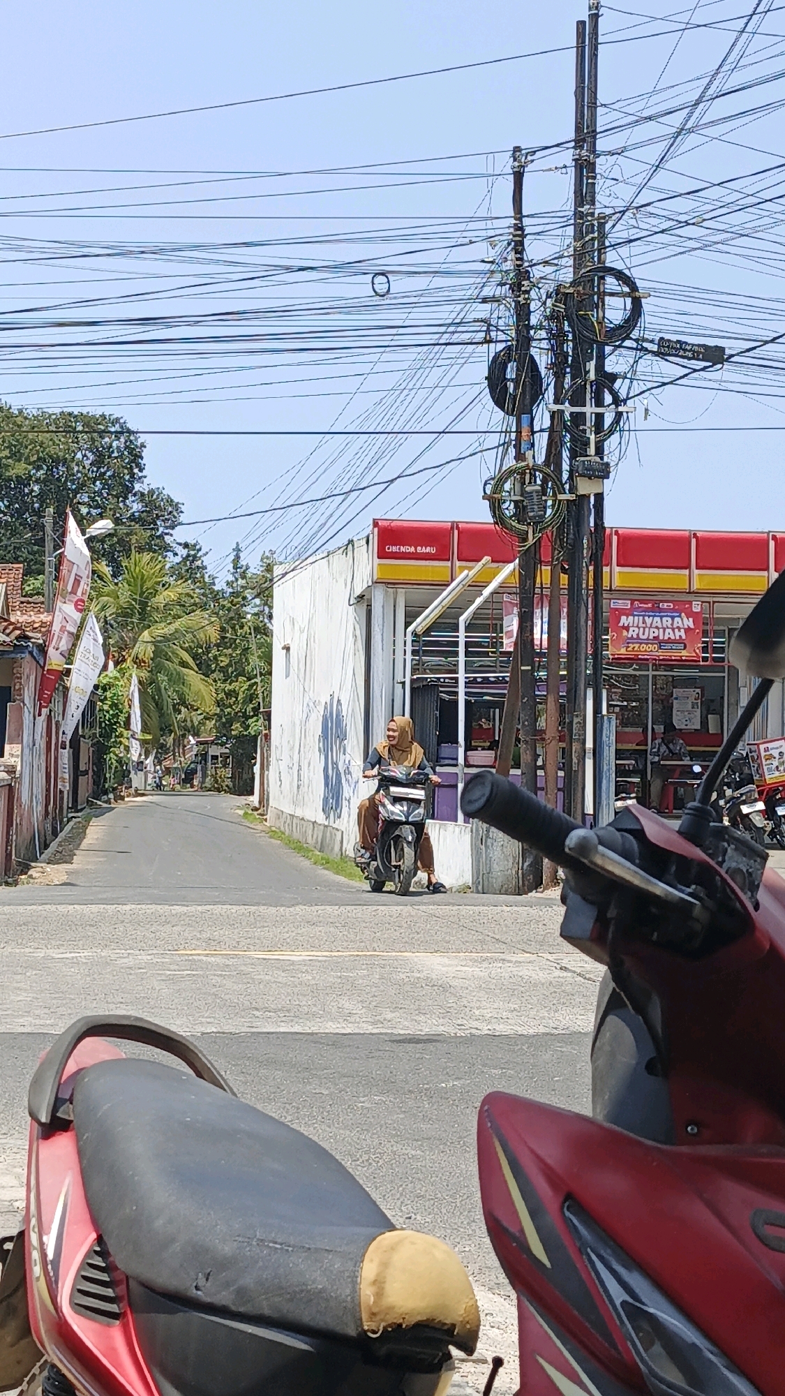 The image depicts a street scene in Cibenda, Indonesia, on a sunny day. In the foreground, the left side of a red motorcycle is visible, focusing on its black seat and rear. Further down the paved street, a woman wearing a hijab and a long brown dress is riding a black motorcycle. She is looking towards the right and smiling. To the right of the street, there is a white building with a red and yellow awning. A sign on the building reads "MILYARAN RUPIAH" with the number "27.000" below it, indicating a promotion or offer. Behind the building, a tall utility pole is laden with a dense network of tangled wires. Palm trees and other foliage are visible on the left side of the street, and other buildings and parked vehicles are present in the background. The overall environment suggests a commercial or residential area within the town.