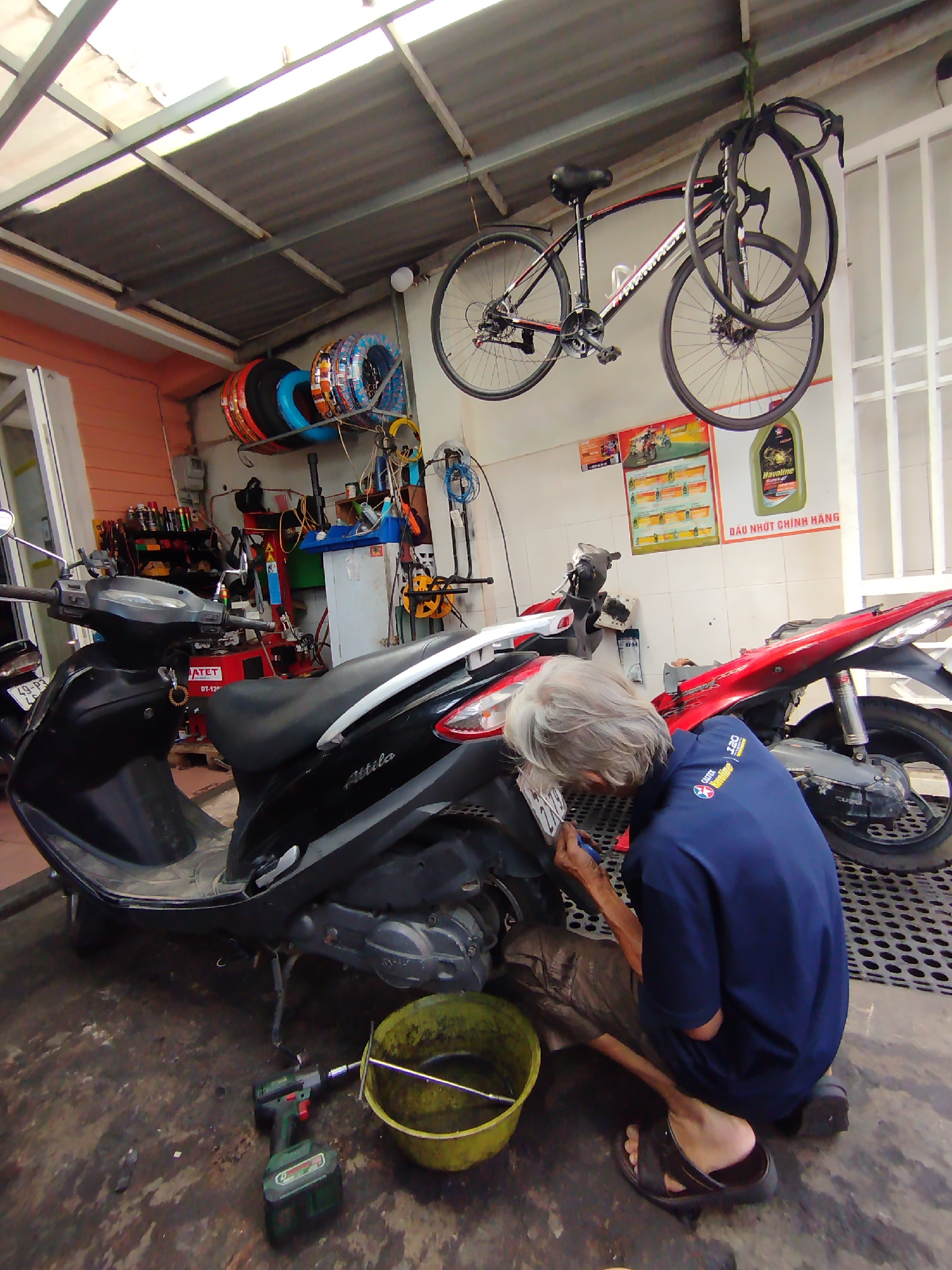 The image captures an elderly person diligently working on a black scooter inside a motorcycle repair shop, likely in Cam Ly - Đà Lạt, Vietnam. The individual, with long grey hair and wearing a blue polo shirt and sandals, is crouched and focused on the rear section of the scooter, possibly performing maintenance or an oil change, indicated by a yellow basin on the floor nearby. A green power drill is also on the ground.
The setting is a busy garage with a translucent corrugated roof supported by metal rafters, allowing natural light to illuminate the space. Tools and equipment are arranged on shelves and hanging on walls. To the left, another black scooter is parked, while a red motorcycle is partially visible behind the worker. A rack displays several colorful stacks of new tires, and a black road bicycle hangs suspended from the ceiling.
Visible text includes "49-P3" on a license plate, "ATET DT-12" on a piece of equipment, "Attila" on the side of the scooter being serviced, and "Havoline 220" and "Havoline" on posters or product labels. A large sign in Vietnamese reads "DẦU NHỚT CHÍNH HÃNG," translating to "Genuine Engine Oil," indicating the shop's offerings. The scene conveys a sense of industrious activity in a typical local repair establishment.