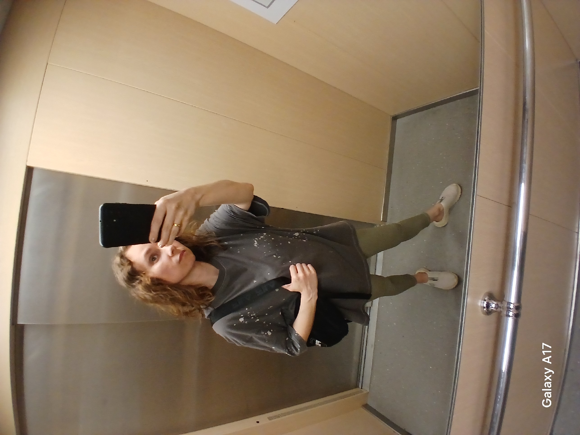 A woman is taking a selfie in what appears to be an elevator. The elevator's interior is paneled in light-colored wood. The woman is wearing a gray t-shirt with white splatter patterns, olive green leggings, and white sneakers. A black shoulder bag hangs from her left shoulder. She is holding a black smartphone in her right hand, taking the picture. The floor of the elevator is gray. A vertical silver handrail is visible on the right side of the frame. The text "Galaxy A17" is printed on the lower right corner of the image. The location is presumed to be somewhere within Yekaterinburg, Russia.