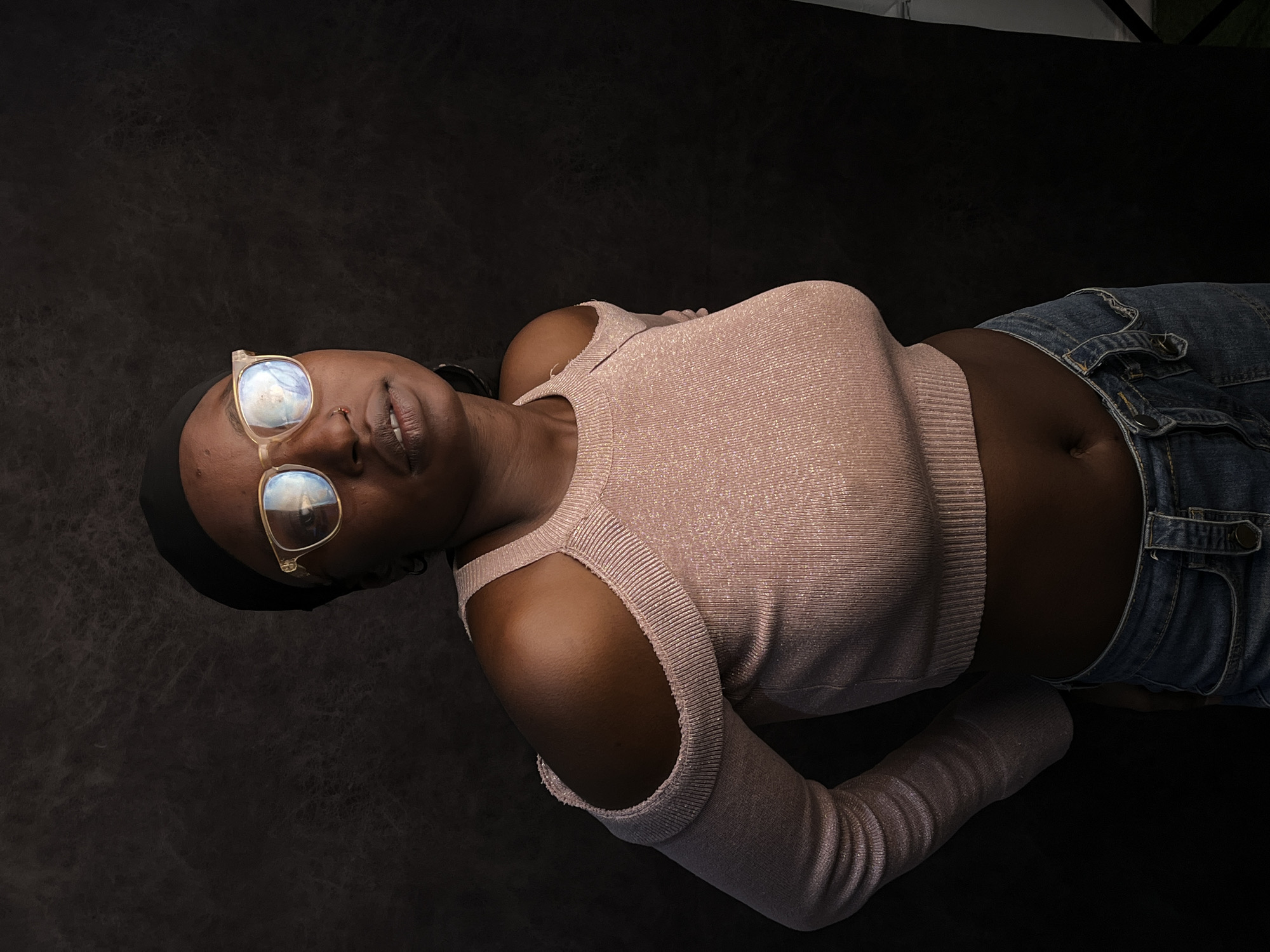 A dark-skinned woman lies on a black surface. She is wearing a pink long-sleeved shirt with shoulder cut-outs and blue jeans. She has a black head covering and clear-framed glasses. There is no indication of the location being Barakin Akawo, Nigeria.