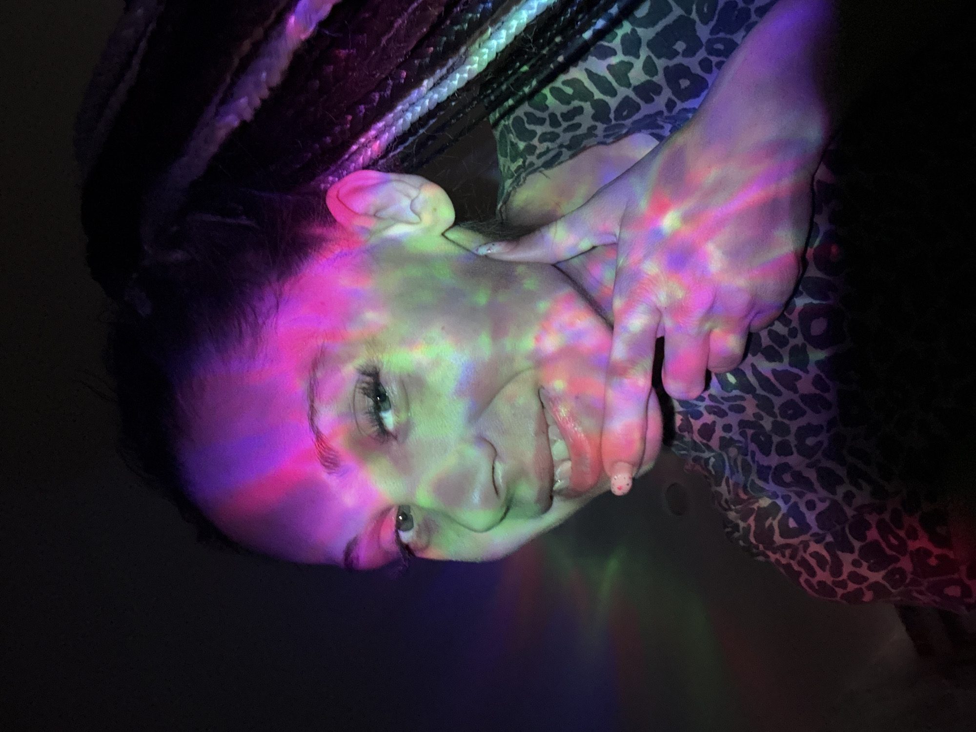 A woman is photographed indoors under colorful, projected lights. The woman has light skin and dark hair, styled in braids. She is wearing a leopard-print top. Her face and hand are illuminated with red, pink, green, and blue light patterns. The background is dark. There are no indications of Rusanovo, Russia in the image.