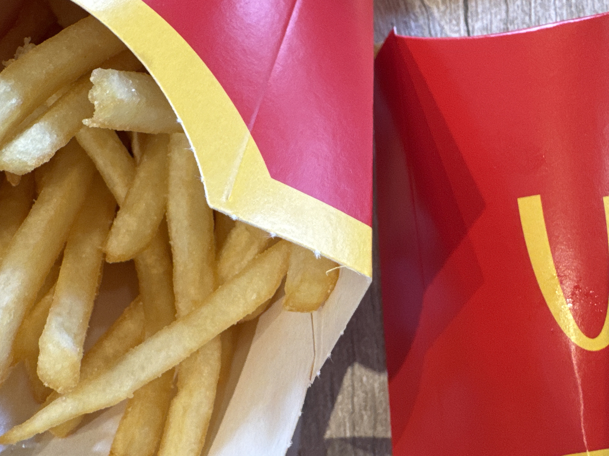The image depicts a close-up of McDonald's french fries in a partially opened red and yellow paper container, resting on a light-colored, textured surface, possibly a wooden table. Another closed McDonald's french fry container is positioned to the right of the open one. The location in Lipomo, Italy is not explicitly stated or visually discernible from the image itself.