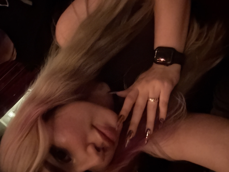 A close-up portrait captures a female individual in an indoor, low-light setting. The subject's head is tilted slightly, and she looks towards the camera. She has long, primarily blonde hair with distinct pink streaks on the viewer's right side (her left). She wears a dark-colored garment. Her left hand is raised, with fingers partially splayed, positioned near her shoulder or upper chest. Her fingernails are long, pointed, and adorned with a metallic or reflective brown/gold polish, some featuring additional patterns. A gold-colored ring with a prominent design is visible on the ring finger of her left hand. A dark-colored smartwatch with a black band is worn on her left wrist. The background is dark and significantly blurred, showing indistinct reddish-brown tones, consistent with warm ambient lighting in an establishment. The specific location within the city of Tel-Aviv, Israel, cannot be determined from the visual information presented in the image.