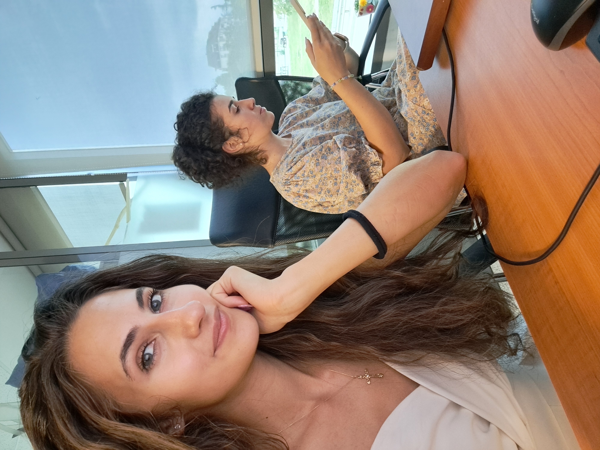 Two women are in an office setting. The woman in the foreground, facing the camera, has long, wavy brown hair and is looking at the camera with a slight smile. She is wearing a white top and a gold necklace with a cross pendant. Her left arm is resting on a wooden desk. The woman in the background is seated at the desk, facing away from the camera, and is looking down at her phone. She has curly dark hair and is wearing a light-colored, floral-print dress. The office has large windows with a view of greenery and buildings outside. A computer mouse and a laptop are visible on the desk. The lighting suggests it is daytime, possibly overcast.