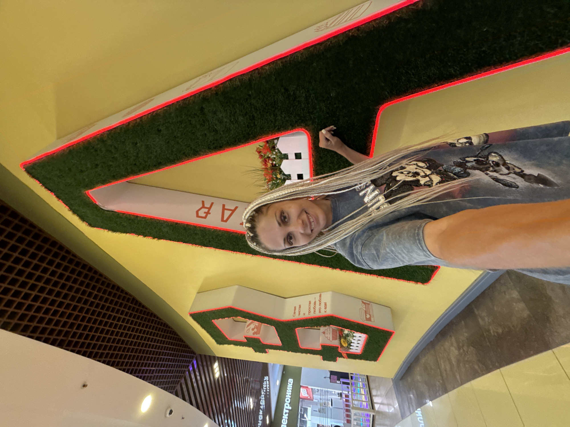 A white woman with blonde braided hair is taking a selfie in front of a large, artificial grass letter "A" with red neon lighting. She is wearing a light blue t-shirt with a graphic print. The letter "A" is mounted on a yellow wall. To the left of the letter "A," there is another artificial grass structure in the shape of the letter "B" with white shelves and red neon lighting. Inside the "B," there are decorative items including a cupcake, flowers in a white picket fence, and a coffee cup with a heart. The background shows a ceiling with a dark brown grid pattern, a white ceiling with lights, and a store called "Elektronika" (Electronics) in the distance. The floor is tiled with yellow and grey tiles. The image was taken in a shopping mall in Rusanovo, Russia.