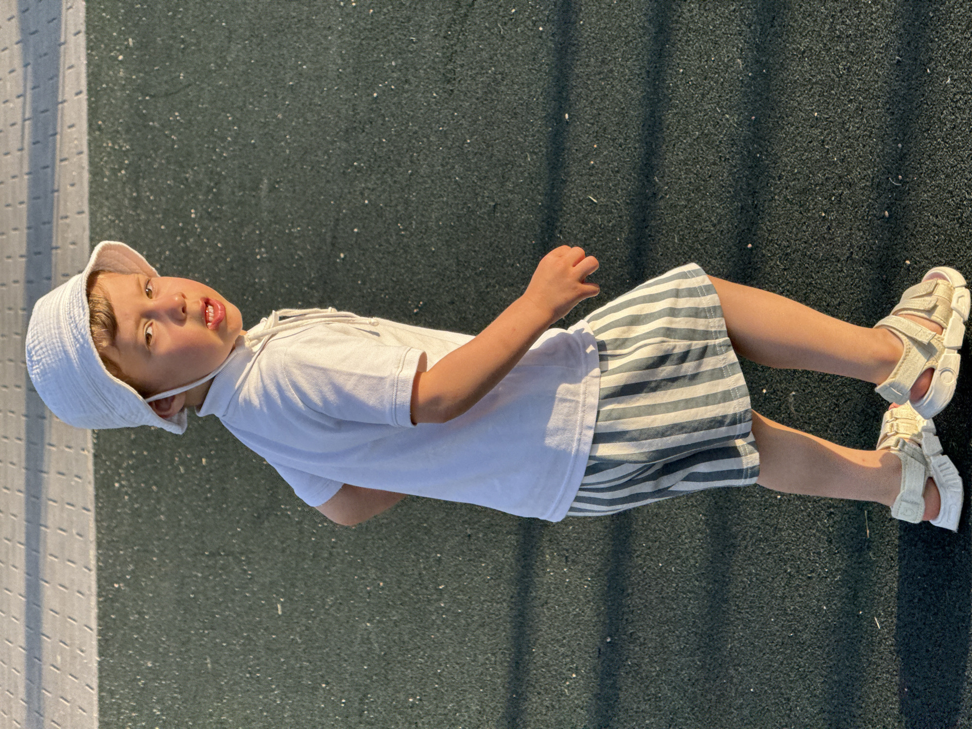 A young boy, wearing a white bucket hat, a white polo shirt, and striped shorts, stands on a dark green, textured surface with a light grey, patterned surface behind him. He is looking back over his right shoulder with a slight frown and his mouth open. The boy is wearing white sandals. The lighting suggests it is either early morning or late afternoon, casting long shadows. The scene appears to be an outdoor recreational area, possibly a playground or sports court.