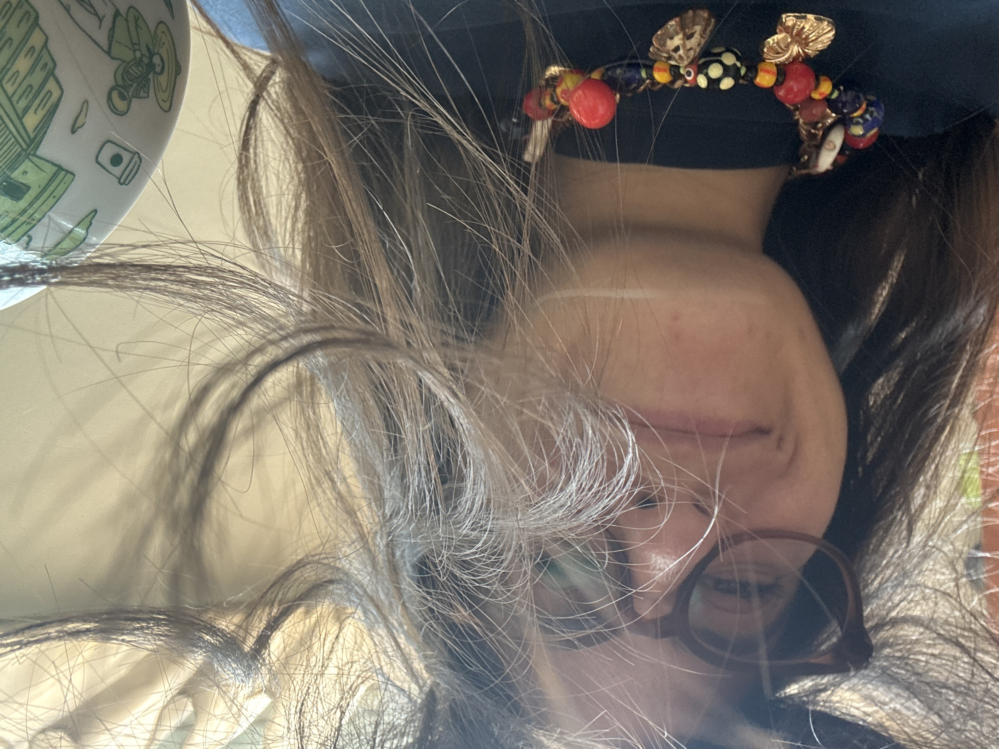 A selfie-style, upside-down image shows a person with dark brown hair streaked with gray, wearing brown-rimmed eyeglasses. The person’s face is partly obscured by hair. They wear a dark shirt with a colorful beaded headband or necklace featuring insects and shells as adornments. A ceramic cup with green cartoon-style drawings is visible in the upper left corner. The location is not identifiable as Puebla, Mexico from the image.
