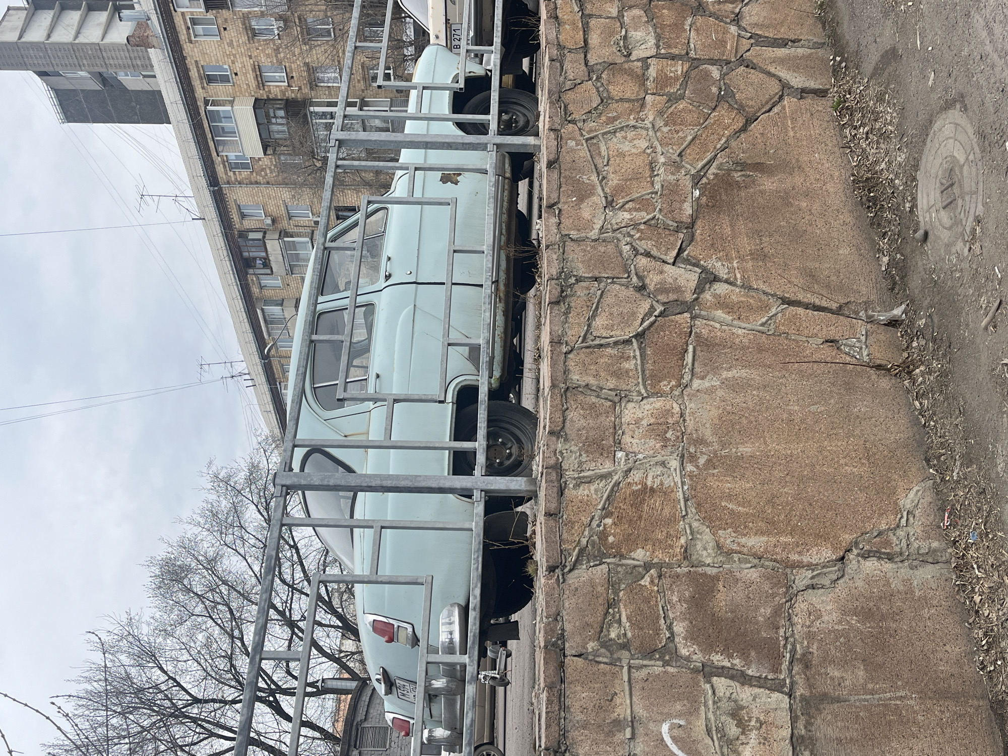 The image shows a light blue GAZ-21 Volga car parked behind a metal fence next to a stone wall. The car has a license plate that appears to be partially obscured. Beyond the car and fence, there is a bare tree and buildings under an overcast sky. The scene is located in an urban area, likely in a city. The exact location in the city of Unknown, Unknown is not discernible. The ground is paved or covered in gravel.