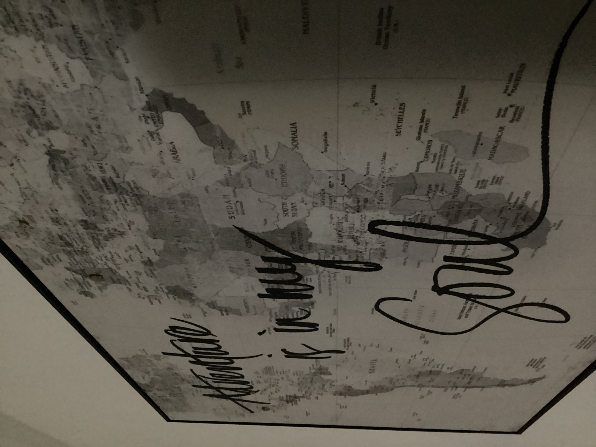 This media file is a photograph of a world map wall decal with a calligraphic overlay. The map itself is detailed, showing continents, countries, oceans, and cities with their names. The calligraphic text is in a cursive font and appears to say "Adventure is in my soul." The map is predominantly grayscale, with some subtle textures suggesting the terrain of the landmasses. The overlay text is in a bold, dark color, making it stand out against the map. The image is taken from an angle, suggesting it is on a wall. The lighting is dim, and there are some reflections on the surface of the decal. There is no indication of people, specific time of day, weather conditions, or emotions conveyed by the image itself. The visible text consists of place names on the map and the overlayed phrase. The context provided states the location as North Port, United States of America, which is not discernible from the image alone.