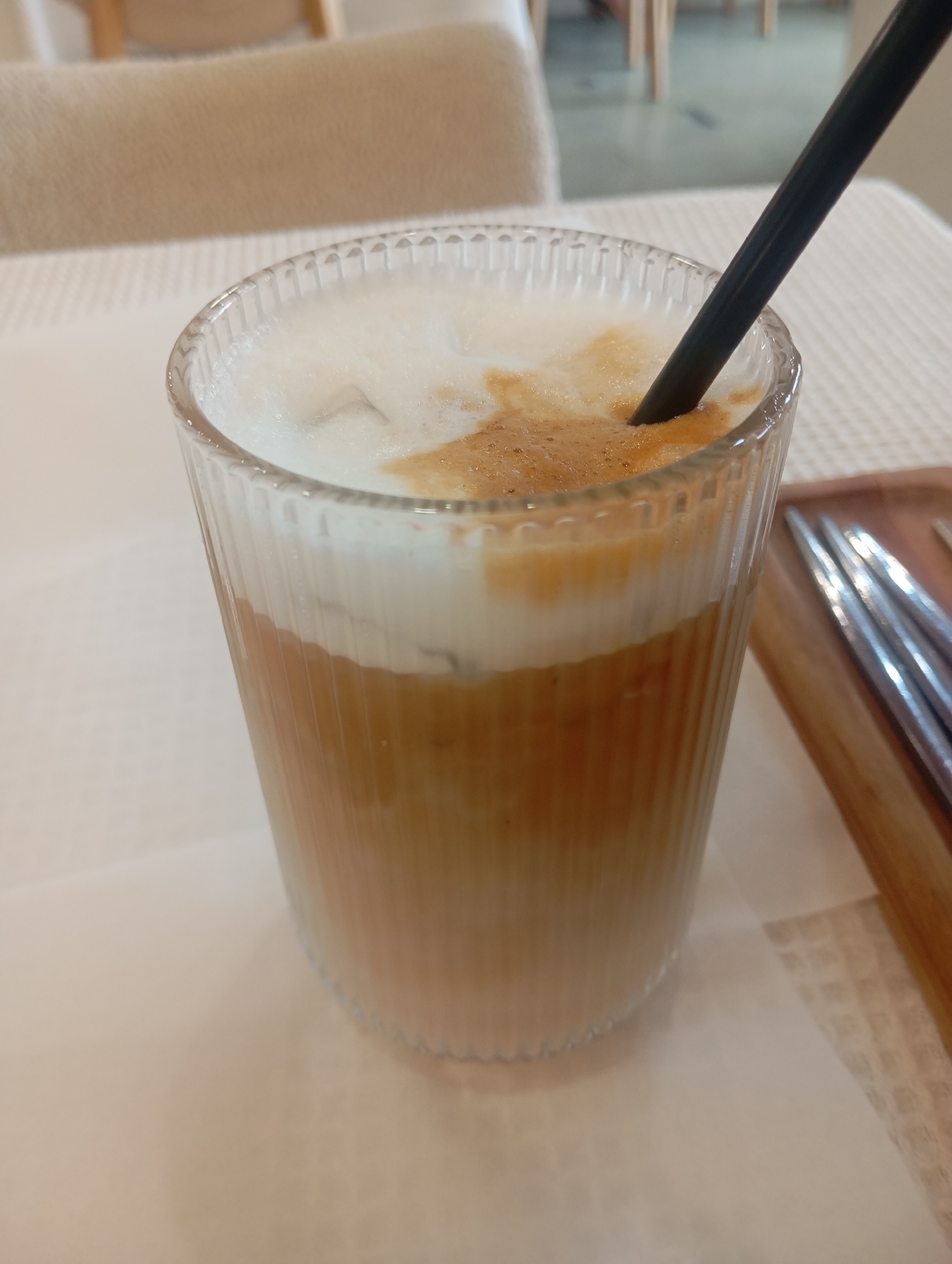 A close-up, eye-level shot showcases a tall, fluted clear glass filled with a layered iced coffee drink, topped with a thick layer of frothy milk and a dark straw inserted into it. The drink itself exhibits a gradient of colors, with a lighter beige at the bottom, transitioning to a richer caramel color in the middle, and finally the white foam on top. The glass is positioned on a textured, light-colored surface, possibly a tablecloth, with a small portion of a wooden serving tray holding metal utensils visible on the right side of the frame. In the background, blurred elements of what appears to be a restaurant or cafe setting are visible, including a light-colored chair and a table. The overall lighting suggests an indoor environment, possibly during daylight hours. There are no people visible in the image. The scene is clean and well-lit, focusing on the beverage. The location of Almaty, Kazakhstan is not discernible from the visual information present in the image.