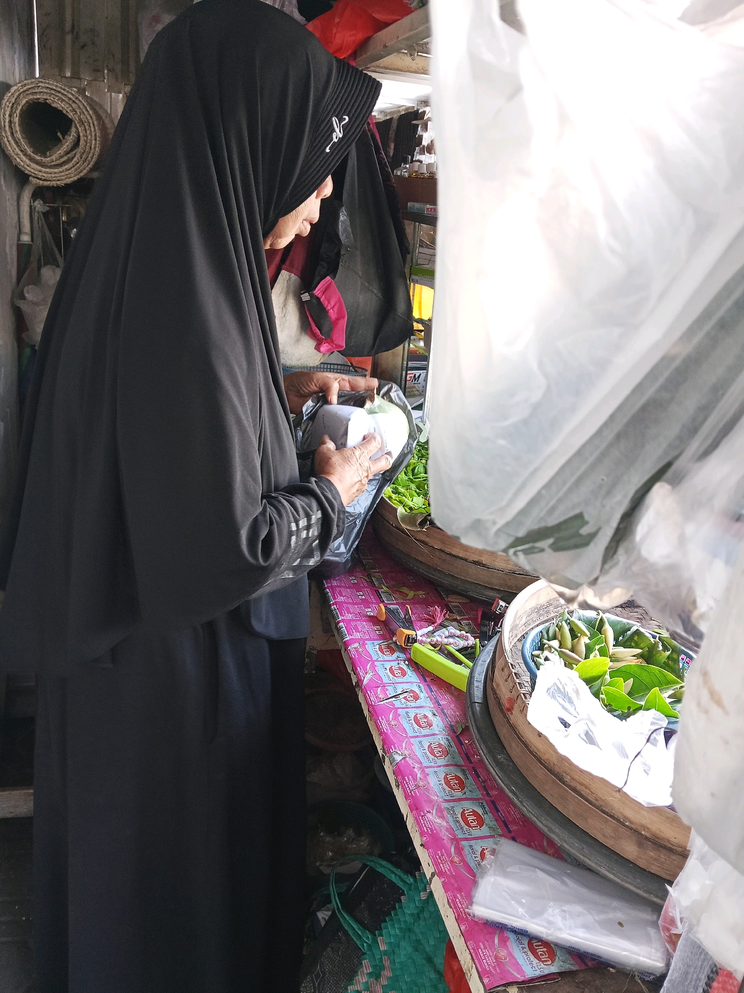 A woman wearing a black hijab is seen from the side, standing in what appears to be a market stall in Wates, Indonesia. She is holding a small white paper bag or packet in her hands, examining it. Her right hand is visible, with fingers bent as she holds the item. Her left hand is mostly obscured by the bag. The woman's face is partially visible, with her profile showing her looking down at the object.
The stall is filled with various items. In the foreground, to the right, is a woven basket filled with green leaves and some lighter colored, possibly conical, objects, perhaps some kind of produce. A clear plastic bag hangs over the front of this basket, obscuring some of the contents. To the left of the basket, on a pink-covered surface, are several items including a bright green stapler-like object, some small white beads or trinkets on a string, and what appear to be several small packets or wrappers with pink and blue designs.
Behind the woman, shelves or racks are visible, laden with more items, some of which are blurry. A rolled-up mat made of natural fibers is leaning against a wall on the far left. The overall environment suggests a small, busy market stall. The lighting is uneven, with some areas brightly lit by what appears to be sunlight coming from the right, and other areas in shadow.