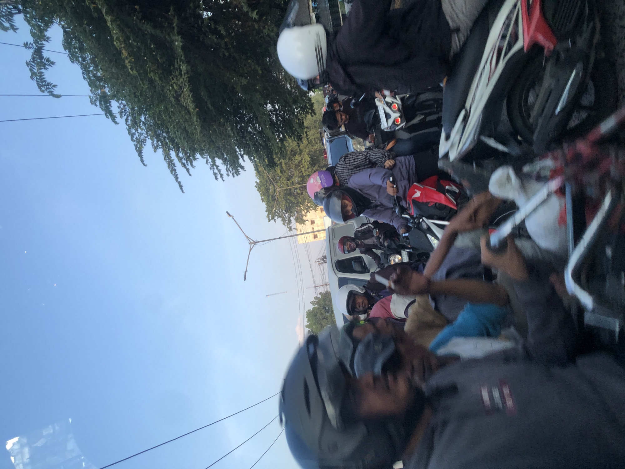 The image depicts a traffic scene in the city of Medan, Indonesia, likely during a congested period. The foreground is dominated by the blurred image of a person wearing a dark helmet, obscuring their face. Behind them, several other individuals on motorcycles are visible, some wearing helmets of various colors, including black, white, and pink. One person in the middle ground wears a purple jacket and a black helmet, while another in the background sports a white helmet and a black jacket. A white van is partially visible to the left, and various trees and power lines are present in the background. The sky is clear and blue, suggesting it is daytime. The overall impression is one of a busy street with many motorcycles navigating through traffic.