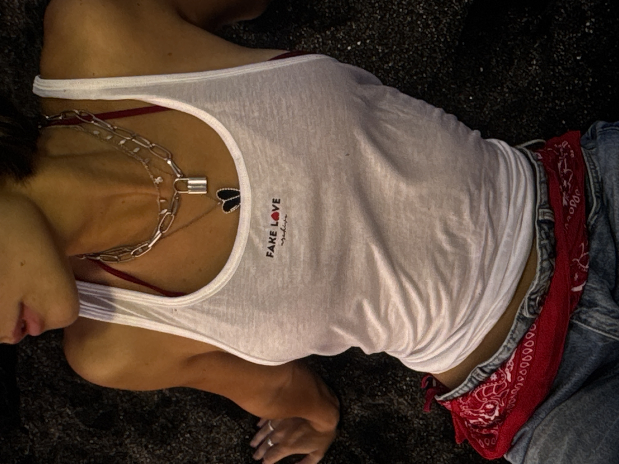 A person is lying down with their upper body visible, wearing a white tank top layered over a red bra. The tank top has a graphic that reads "FAKE LOVE" with a red heart. They are also wearing multiple necklaces, including a chunky silver chain and a pendant shaped like a heart with black accents. Around their waist, they have a red bandana tied and are wearing denim jeans. The person is lying on a dark, textured surface that appears to be sand or gravel. Their right hand, resting on the surface, has a ring on it. The overall impression is a casual, relaxed pose. This scene could be located on a beach in Kuta Selatan, Indonesia, given the textured ground and relaxed attire.