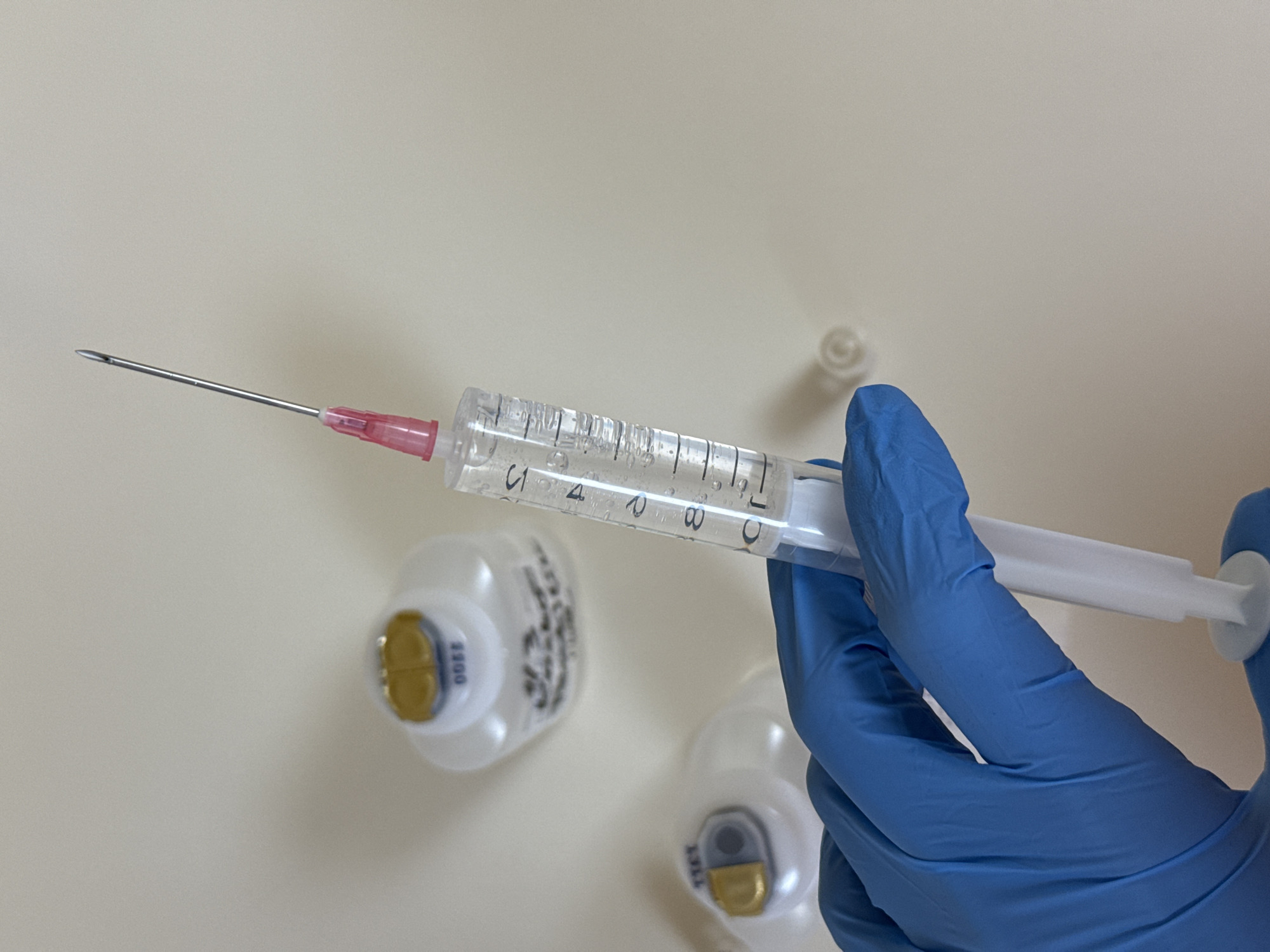 A gloved hand holds a syringe filled with a clear liquid, with the needle pointing to the left. Two medicine vials are on a light surface in the background, along with a white cap. The location in Pleven, Bulgaria, cannot be determined from the image.