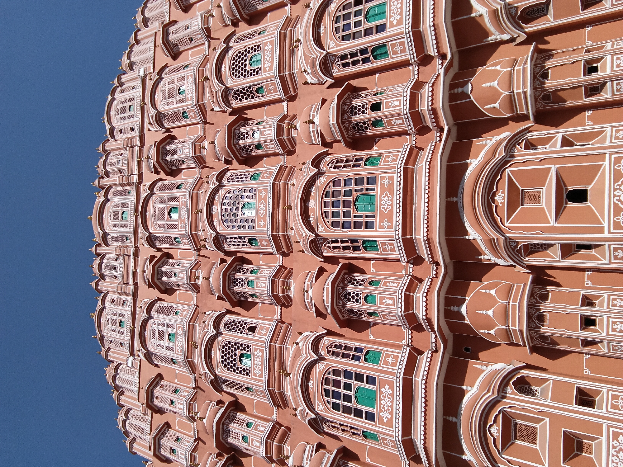 The Hawa Mahal, a palace in Jaipur, India, is depicted in a low-angle shot against a clear blue sky. The facade is made up of a honeycombed structure, featuring numerous small windows with ornate designs. The building is primarily reddish-brown, with white decorative outlines highlighting the architecture. The windows appear to have green or darkened interiors, possibly indicating stained glass or shadowed openings. The palace's exterior exhibits a pattern of projecting bays, creating a rhythmic and layered effect. The angle of the shot emphasizes the height and intricate detail of the building.
