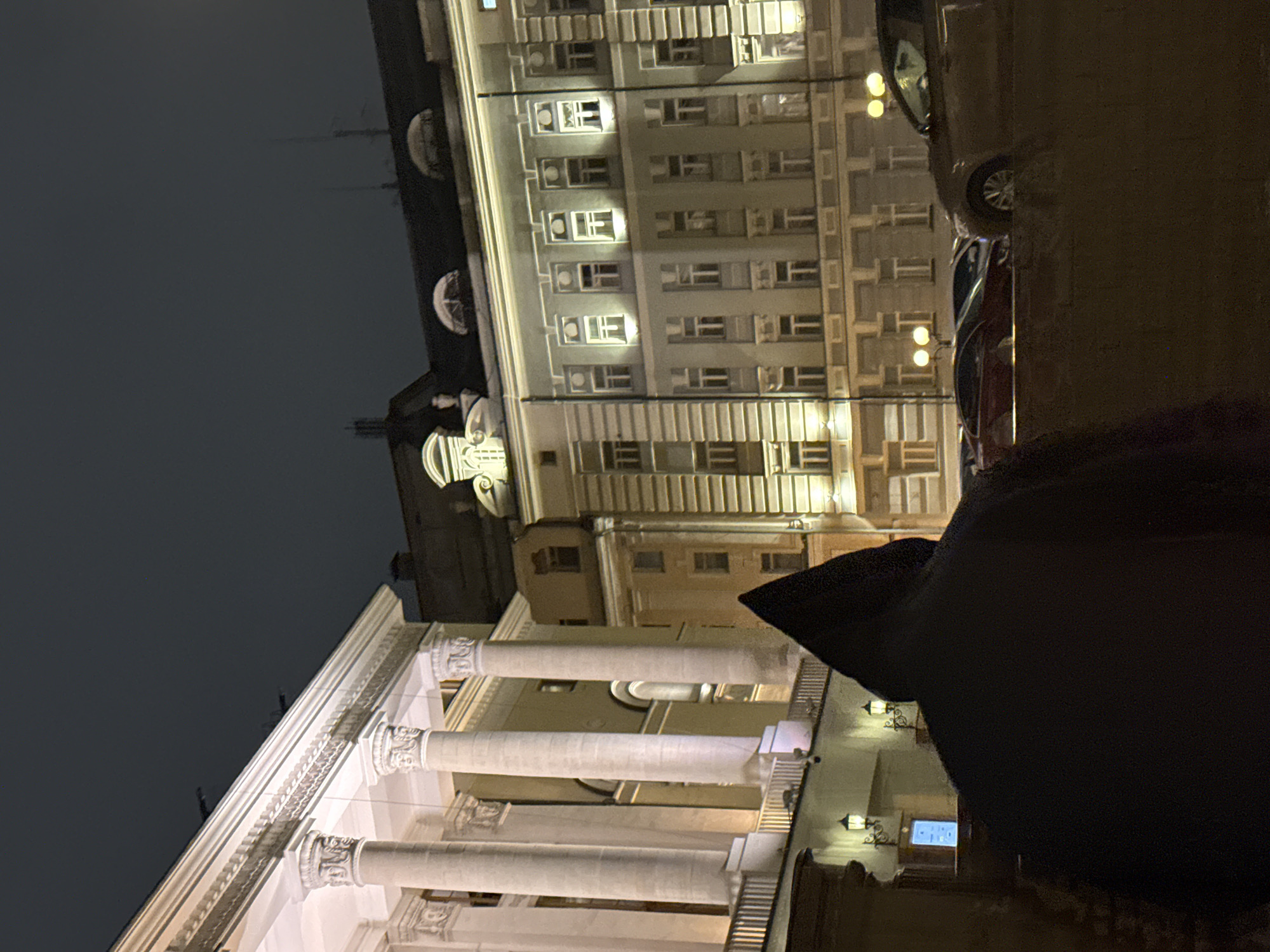 The image shows an exterior view of the Bolshoi Theatre in Moscow, Russia, at night. The theater's facade, with its columns and architectural details, is illuminated. A tall building is next to the theater. The image appears to be taken from street level and includes a portion of what appears to be a person's head and part of a car. The sky is dark, suggesting it is evening.
