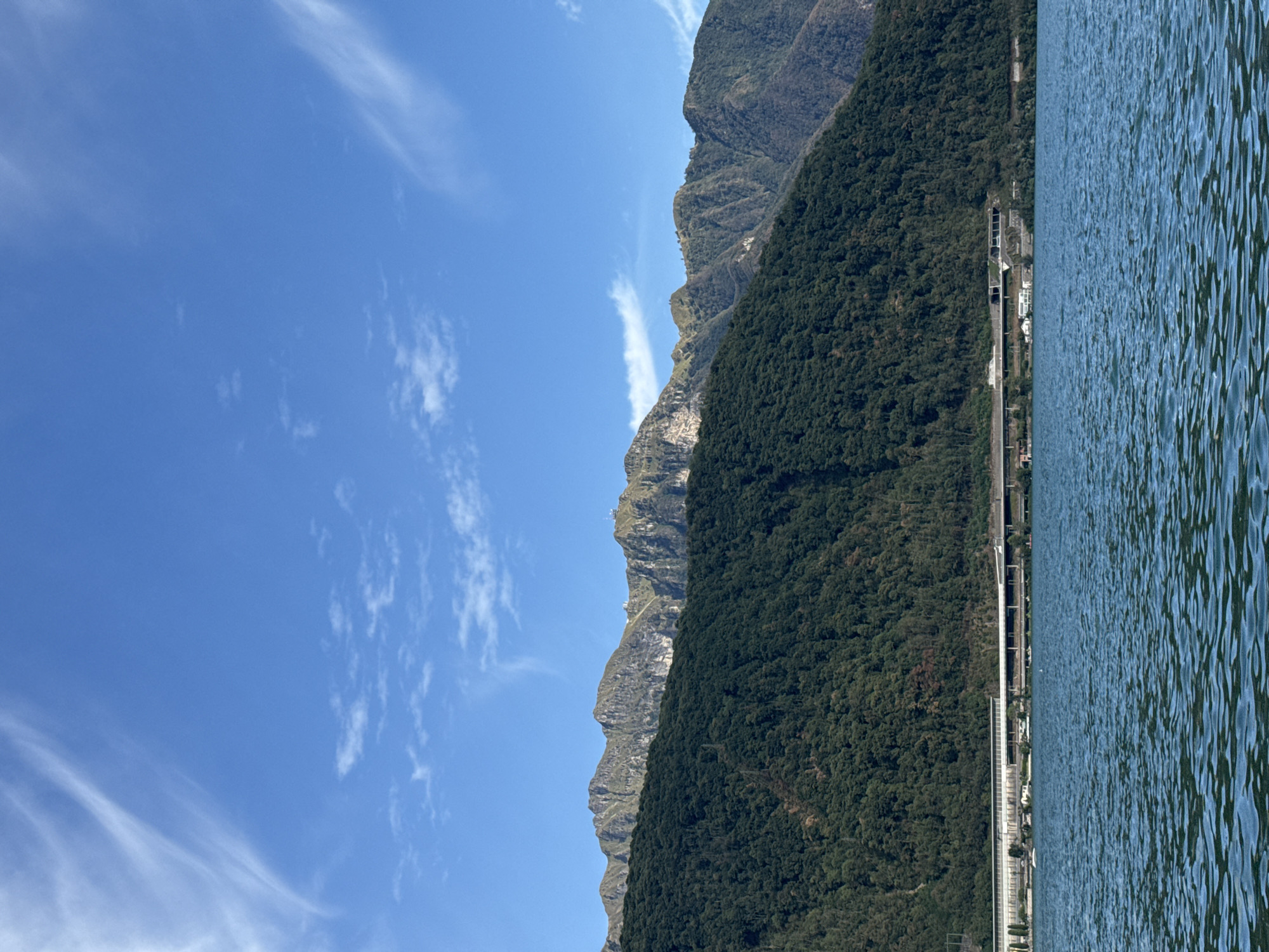 The image shows a view of Lake Lugano in Melide, Switzerland, from the water. In the foreground, the deep blue water of the lake is visible with gentle ripples. Beyond the water, a treed mountainside dominates the middle ground, covered in dense green foliage. Along the base of the mountain, a man-made structure, likely a highway or railway line with retaining walls and possibly tunnels, runs horizontally. In the background, rugged, rocky mountains rise, their peaks reaching towards a bright blue sky scattered with wispy white clouds. The overall scene depicts a natural landscape integrated with human infrastructure.