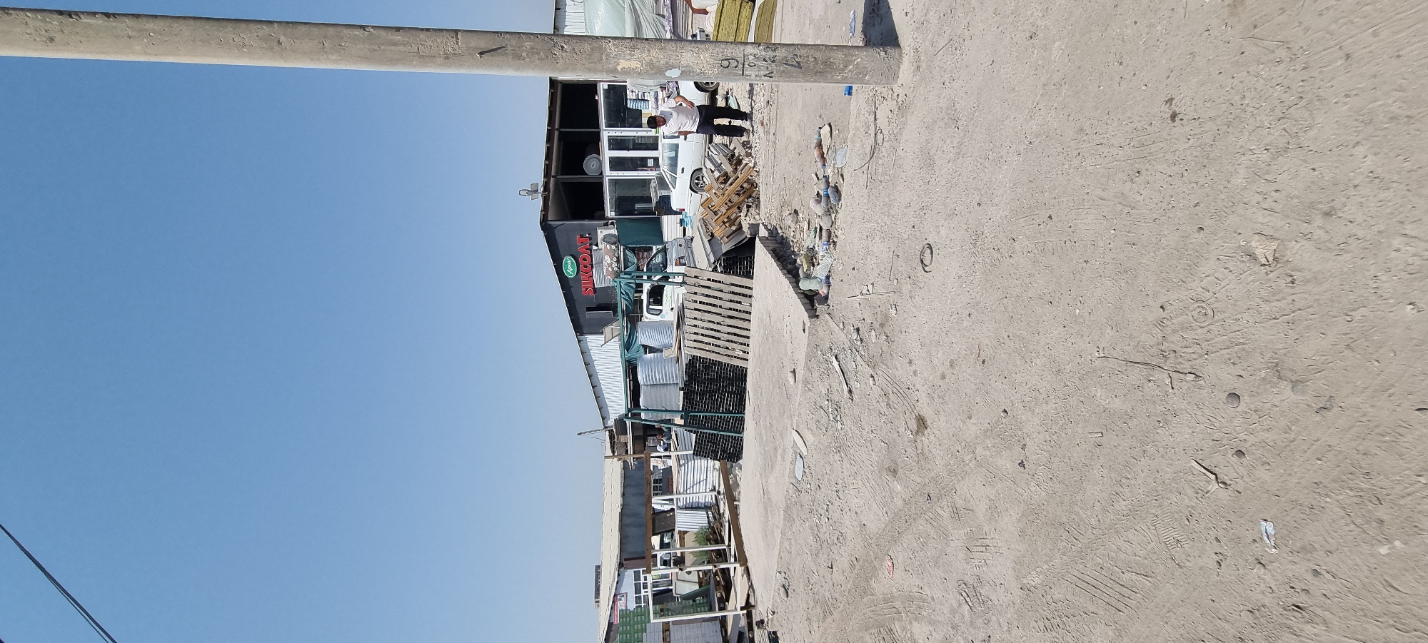 The image depicts an outdoor scene in Bokhtar, Tajikistan, likely a construction material supply yard or a similar commercial establishment. In the background, a building with "SILKCOAT." branding is visible. The foreground is a dusty, unpaved area with various construction materials, including stacks of metal framing, wooden pallets, and large cylindrical objects that appear to be insulation or storage containers. Several vehicles are parked near the building, including a white sedan and two small white trucks. A man in a white t-shirt and dark pants is standing in the middle right of the frame, facing away from the camera and towards the building. The ground is littered with debris, including plastic bottles and other refuse. A utility pole stands prominently on the right side of the image. The sky is clear and blue, suggesting a sunny day.