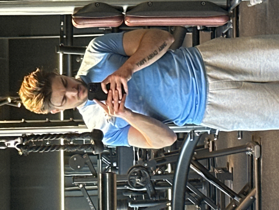 An image captured in a gymnasium in Jestetten, Germany, depicts a male subject taking a mirror selfie. The subject, a Caucasian male with light brown hair styled upwards and a short beard, stands facing forward, looking down at a dark-colored smartphone held in both hands. He is wearing a light blue short-sleeved t-shirt and light gray sweatpants. Script tattooing is visible on his inner left forearm. The background, visible as a reflection in the mirror, shows various gym equipment, including a cable machine with a rope attachment, weight stacks, and upholstered pads on other machines. Overhead spotlights are also reflected, illuminating the scene.