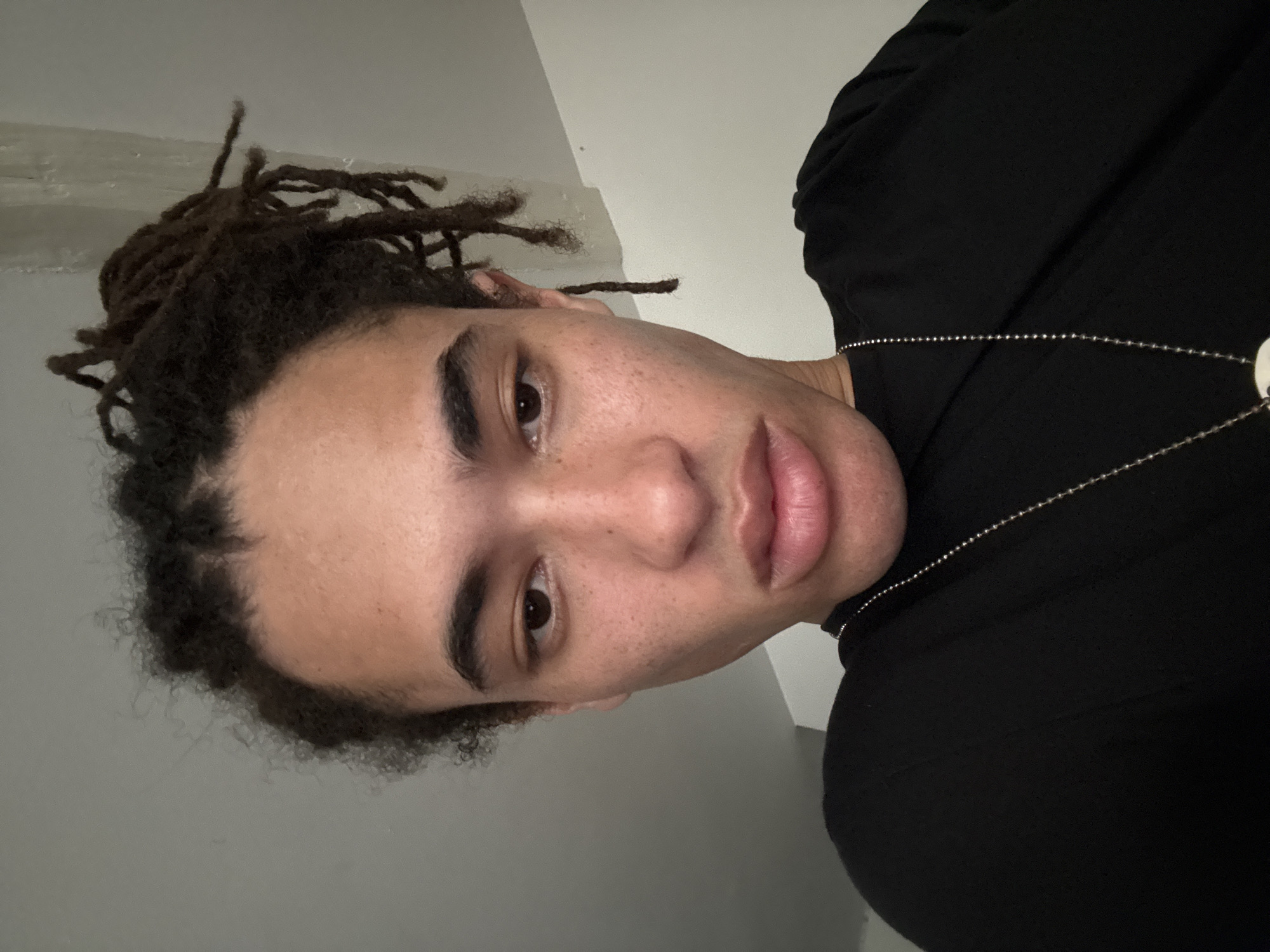 The image is a close-up, low-angle selfie of a young Black individual with dreadlocks and light brown skin with freckles. The person is wearing a black t-shirt and a silver chain necklace. The background is a plain, light-colored wall with some slight texture. The lighting suggests an indoor environment, possibly a room. There are no discernible landmarks or details indicating a specific location in Paris, France. The individual's expression is neutral, and their gaze is directed downwards. The focus is primarily on the person's face and upper torso.