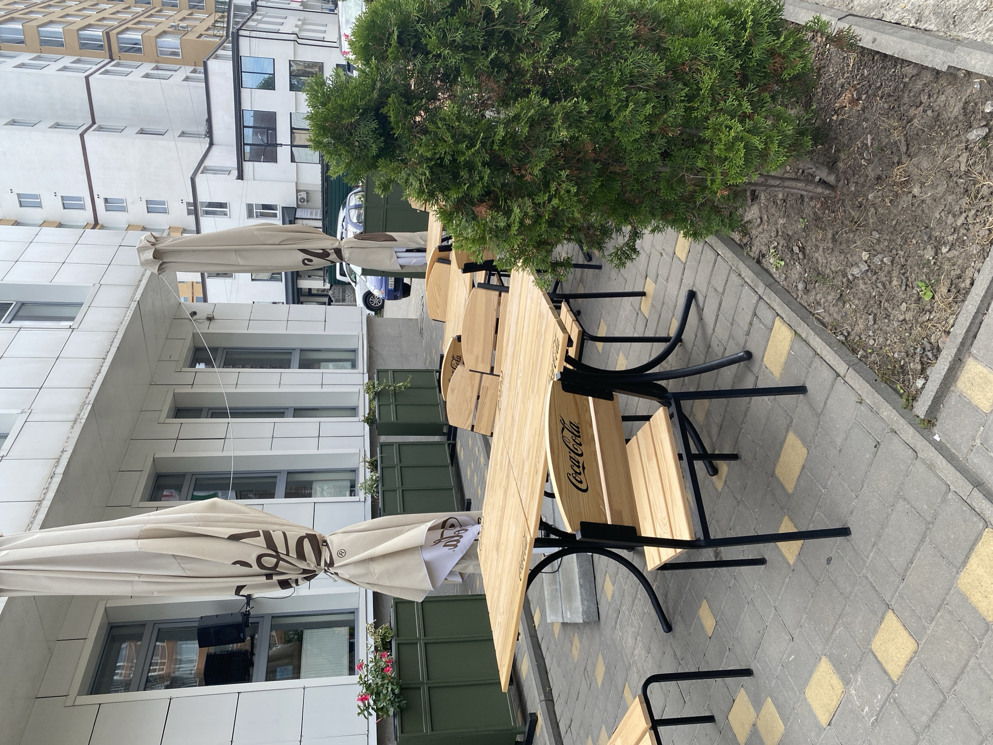 The image depicts an outdoor seating area for a cafe or restaurant located on the street in Vinnytsia, Ukraine. The scene features several wooden tables and chairs, some of which are branded with the Coca-Cola logo. Large, folded umbrellas are positioned behind the tables, suggesting that this area is used for outdoor dining. Planters with flowers and greenery are visible, contributing to the ambiance. In the background, a white building with modern architecture and several windows stands, with other buildings and a street visible further in the distance. The ground is paved with interlocking stones in a checkered pattern of grey and yellow. A mature evergreen tree is planted in a raised bed in the foreground, adding a natural element to the urban setting. The overall impression is that of a clean and well-maintained outdoor dining space.