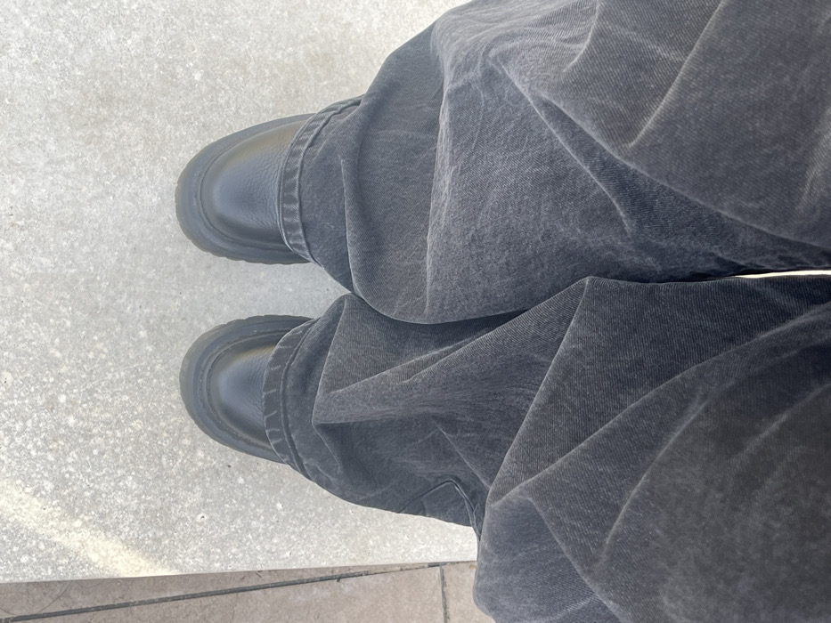 The image displays a downward, first-person perspective focusing on the feet and lower legs of an individual standing on a light-colored, granular paved surface. The individual is wearing black, rounded-toe shoes or boots with thick, textured soles. Their legs are clad in loose-fitting, dark grey denim trousers, which show a faded, stone-washed appearance and numerous folds. A narrow, lighter-colored vertical strip is visible along the far left edge of the frame, indicating an adjacent architectural element or surface. No other individuals, distinct objects, or specific actions are present. The scene is an outdoor paved area in Općina Jasenice, Croatia.