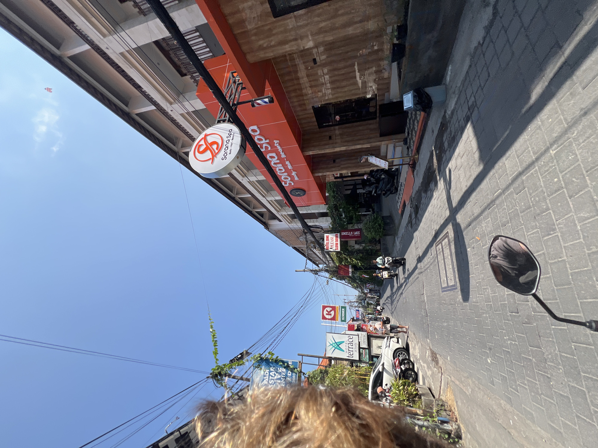An eye-level, medium shot captures a street scene in Kuta, Indonesia, on a sunny day. The street is paved with grey interlocking bricks. On the right side of the street, a two-story building displays the "Sorana Spa" sign prominently, with the words "Body - Hair - Beauty" underneath. The building has an orange facade with brown wooden paneling. To the left of the spa, other businesses and signs are visible, including "Hotel Terrace" and a red K symbol for "Körner." Several motorcycles and a white car are parked along the left side of the street. In the foreground, the rider's left arm and shoulder are visible, along with the handlebar and rear-view mirror of a motorcycle. Two people are seen riding motorcycles further down the street, and another person is walking. Power lines are strung across the bright blue sky above.