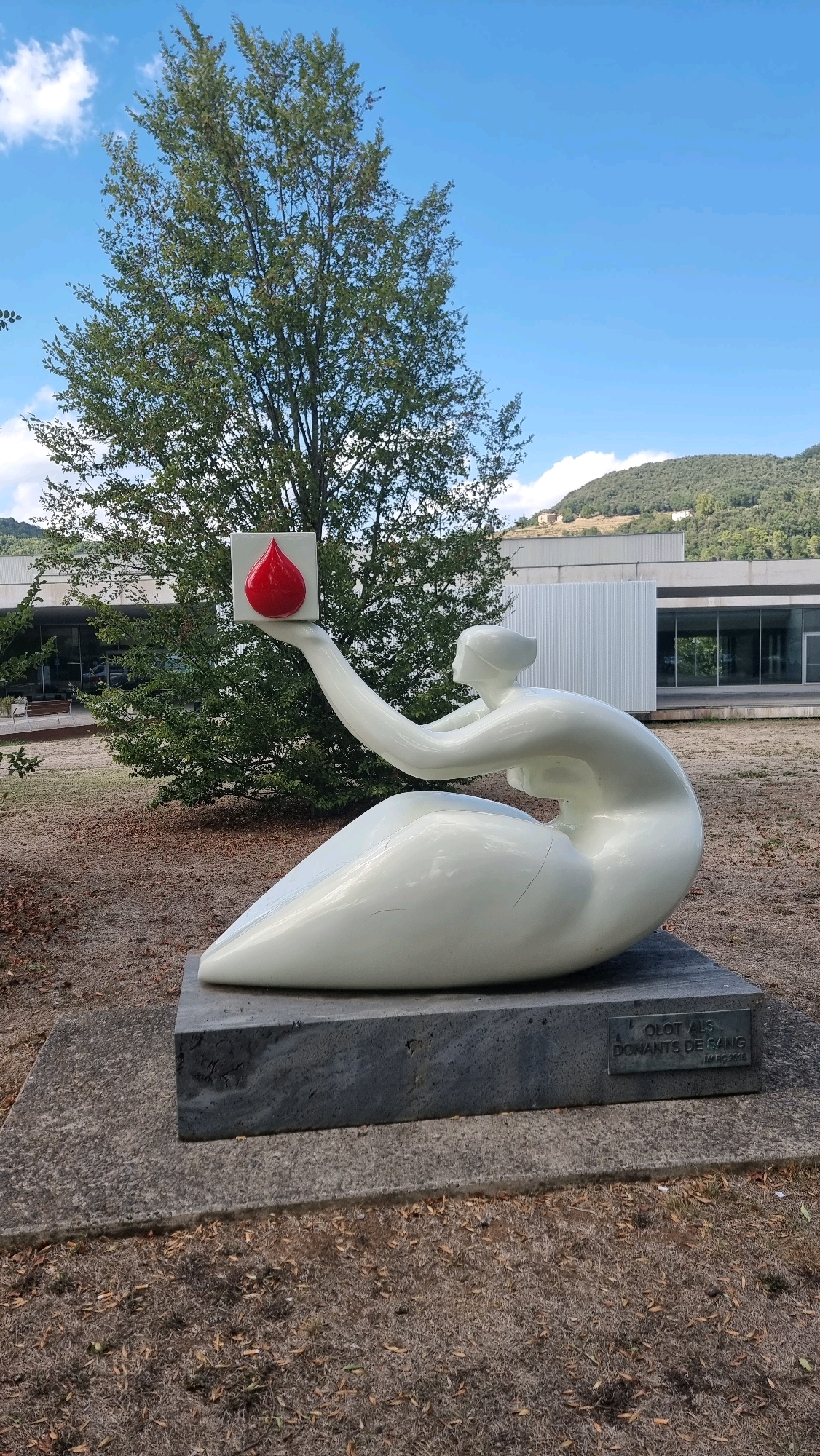 A white, abstract sculpture of a seated figure holding a red blood drop in a white square is displayed on a granite pedestal outdoors. The sculpture is situated in front of a modern building with large glass windows, surrounded by dry grass and a large tree. A plaque on the pedestal reads "OLOT ALS DONANTS DE SANG" and "MARC 2015." The scene is set against a backdrop of a blue sky with some clouds and a green, hilly landscape in the distance. The sculpture represents a tribute to blood donors.