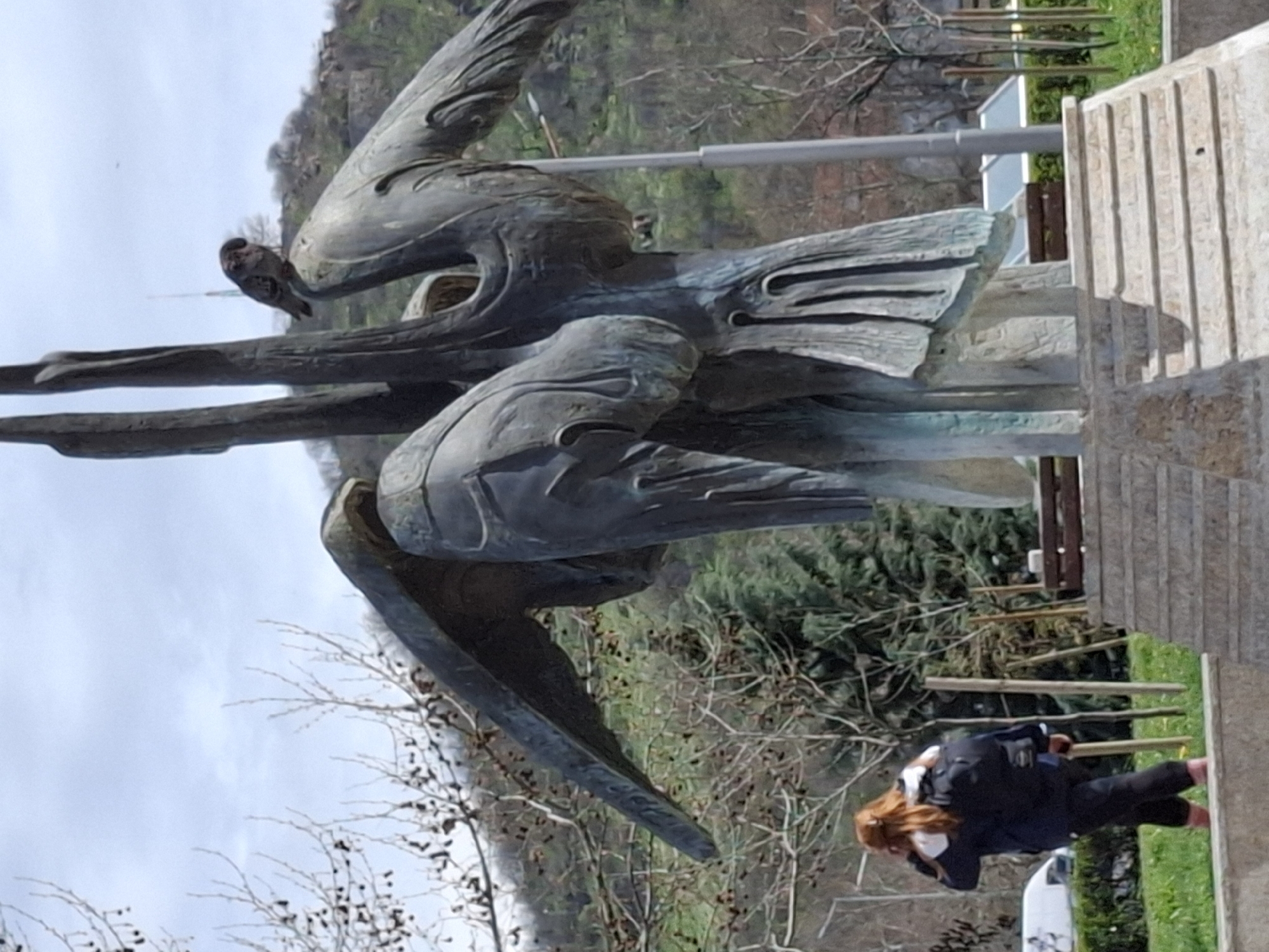 The image shows a bronze sculpture of a winged figure with a bird perched on one of the arms. The sculpture stands on a stone base. Behind the sculpture is a hillside with trees. In the foreground to the right, a woman with red hair is crouched down, appearing to take a photo with her phone. She is wearing a black jacket and pants with a white face mask. Part of a white vehicle is visible in the background near the woman. The location is most likely in the city of Plovdiv, Bulgaria, based on the nature of the sculpture and general environment.