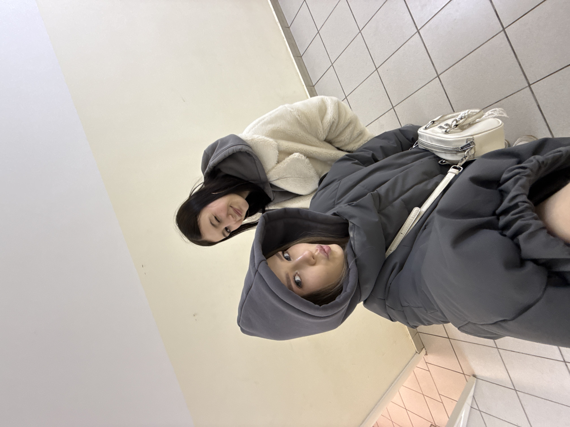The image features two young women captured in an indoor setting, viewed from a slightly low angle.
In the foreground, a young woman with dark hair is seen, wearing a dark grey hooded puffer jacket with the hood up. She looks directly at the camera with a neutral expression, her lips subtly parted in a slight pout.
Behind and to the right of her, a second young woman is visible. She has dark hair and wears a cream or off-white fuzzy jacket, likely a sherpa material, which appears to have dark grey trim around the hood. She offers a gentle, faint smile while looking towards the camera.
A small, light-colored handbag, possibly white or cream, with a silver chain detail and a zipper, rests on the dark grey jacket of the front woman.
The background consists of plain walls and a tiled floor. The wall on the left side of the frame is a uniform light beige or cream color. The floor beneath the subjects is covered in light-colored, possibly white or light grey, square tiles, some of which display distinct diagonal grid patterns. The warm clothing worn by both subjects suggests the environment might be cool. There is no visible text in the image. The overall mood appears casual and relaxed, indicative of a posed photograph in a public or private indoor space within Chaikovsky, Russia.