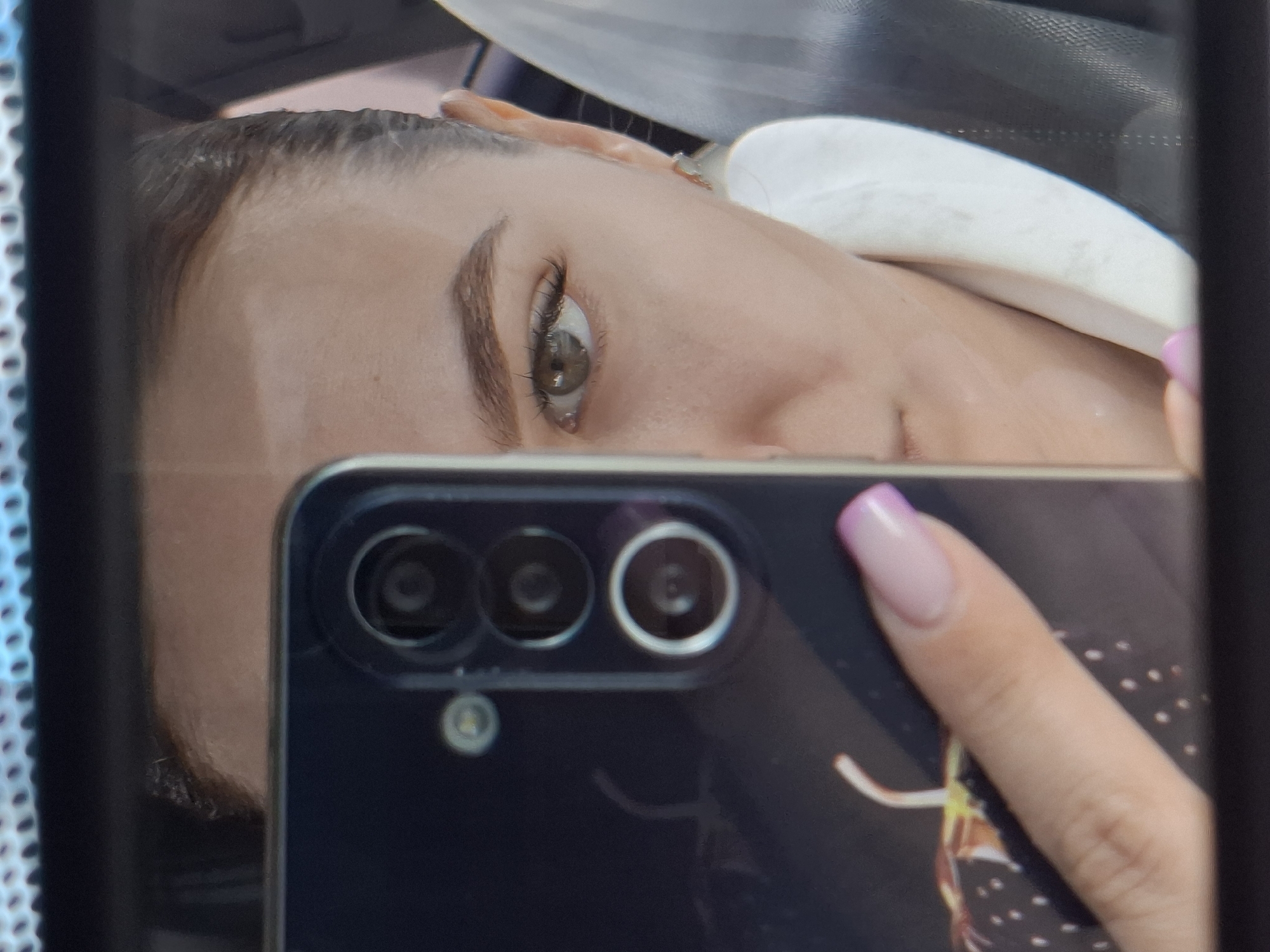 A reflective selfie-style photograph shows a fair-skinned woman partially obscured by a black smartphone held in her right hand. The phone, featuring a triple-lens camera system and a faint design on its back, is positioned in the foreground, partially blocking the woman's face. The woman's left eye, eyebrow, and part of her forehead are visible. Her hair is dark, and she appears to be wearing a white headrest or pillow beneath her head. Her fingernails are manicured with a pink and white design. The background is blurry and indistinct, suggesting an interior setting. There are no discernible elements in the image to place it in the city of Chaikovsky, Russia.