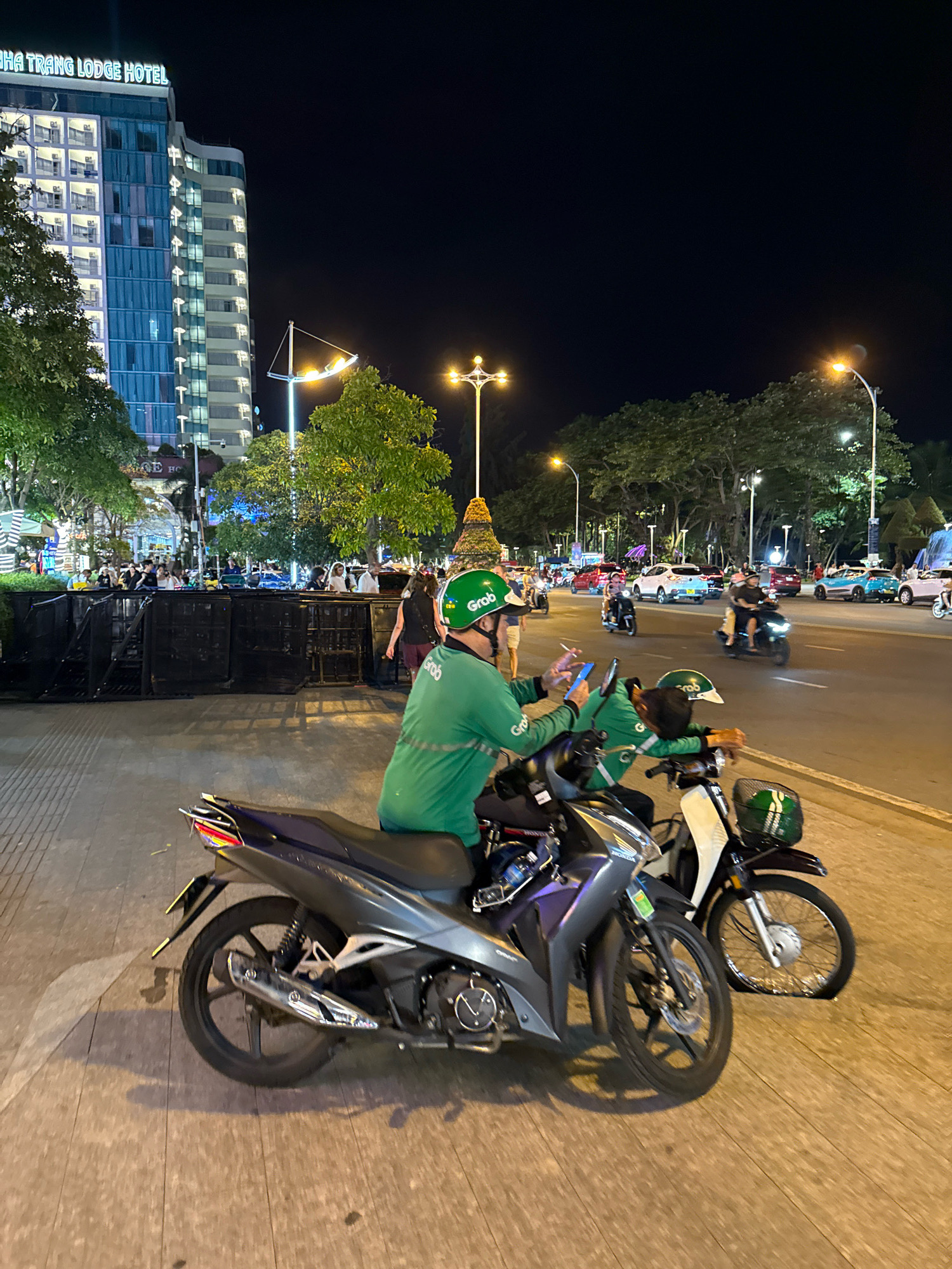 The image depicts a nighttime scene on a busy street in Nha Trang, Vietnam. In the foreground, two Grab delivery drivers are present. One driver, wearing a green uniform and helmet with the Grab logo, is seated on a dark-colored motorcycle and appears to be looking at his phone. The other Grab driver, also in a green uniform and helmet, is leaning forward on his light-colored motorcycle, with his head resting on the handlebars, suggesting he may be fatigued or taking a break. Both motorcycles are parked on a paved sidewalk adjacent to the street.
The background reveals a modern, multi-story building with illuminated windows, identified by a sign as the "NHA TRANG LODGE HOTEL". Beyond the hotel, a street lined with trees and streetlights is visible, with several cars and motorcycles in motion, indicating ongoing traffic. People are also seen walking along the sidewalk and across the street, contributing to the lively atmosphere of the urban environment. The scene is illuminated by artificial lighting from the hotel, streetlights, and vehicle headlights, creating a typical urban nighttime ambiance.