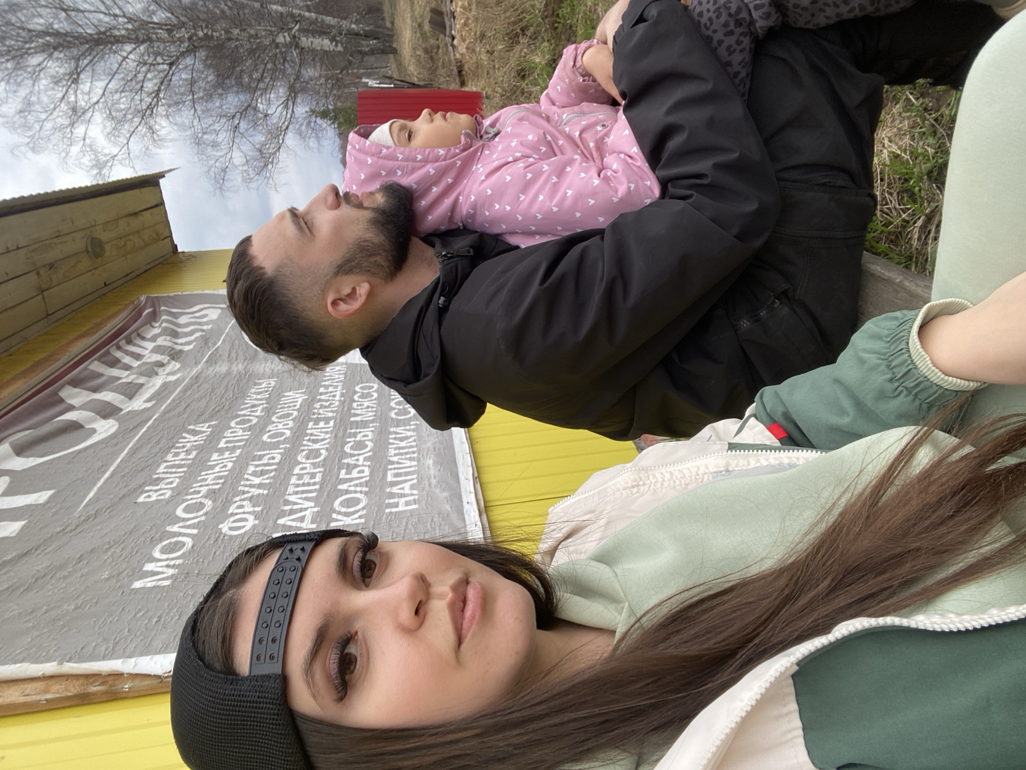A photo taken in Zhigalki, Russia, shows three individuals: a woman with long brown hair wearing a black cap and a light green and white jacket, a man with a beard wearing a black jacket, and a child in a pink jacket lying on the man's chest. The woman is in the foreground, facing the camera. Behind them is a building with a sign written in Russian advertising products, including baked goods, dairy products, fruits, vegetables, sausages, meat, and drinks. The background suggests an outdoor environment with trees and vegetation.