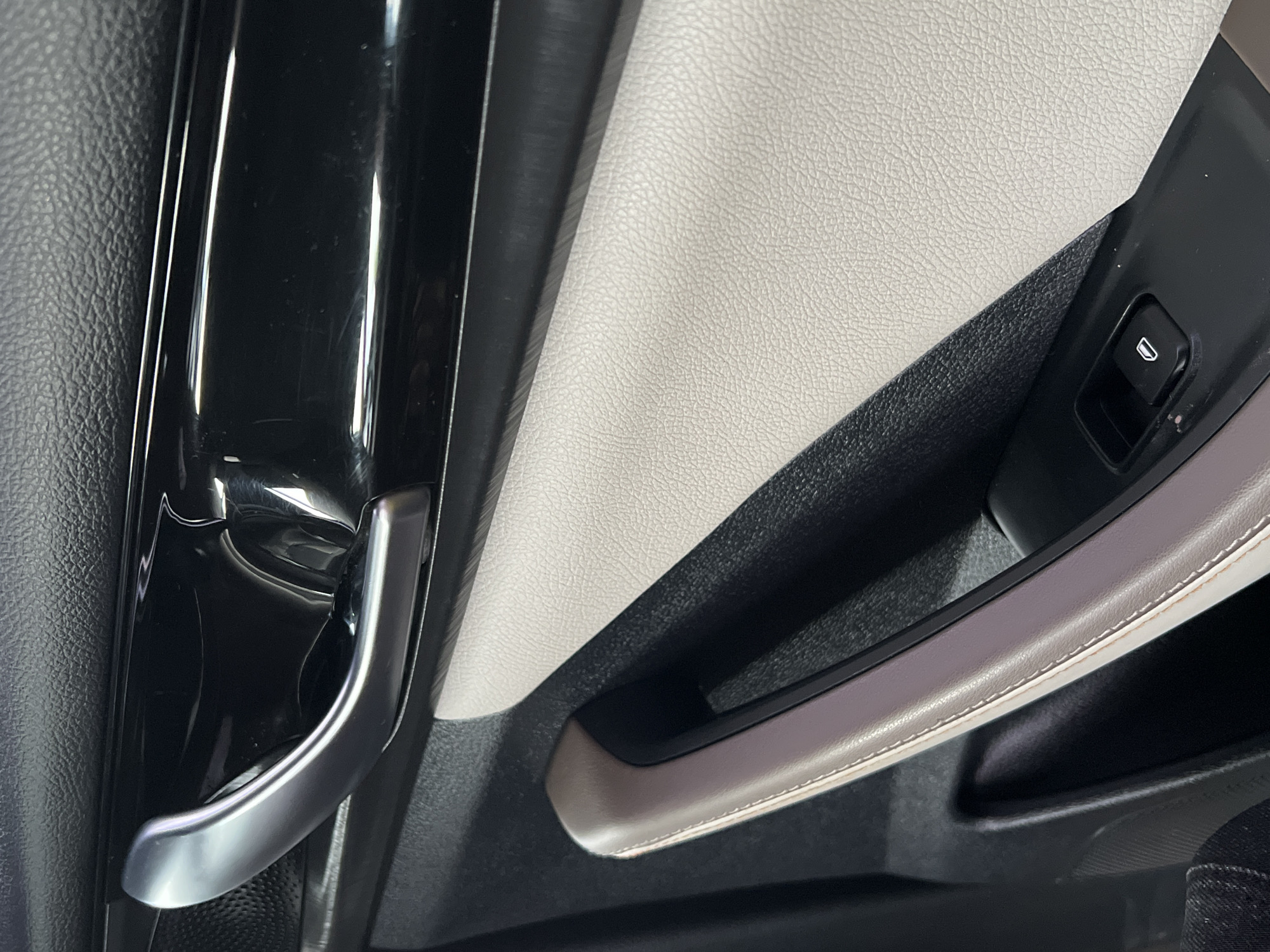 The image displays the interior of a vehicle, focusing on the driver's side door panel. Prominently featured is a chrome door handle with a black glossy trim piece above it. Below the handle, a light-colored textured surface, likely leather or a similar material, forms the armrest. A black window control button with a white icon depicting a lowered window is visible on the armrest. The environment appears to be indoors, with controlled lighting casting reflections on the glossy surfaces. No specific details are present to indicate a location in Yekaterinburg, Russia, or any people or actions within the scene.