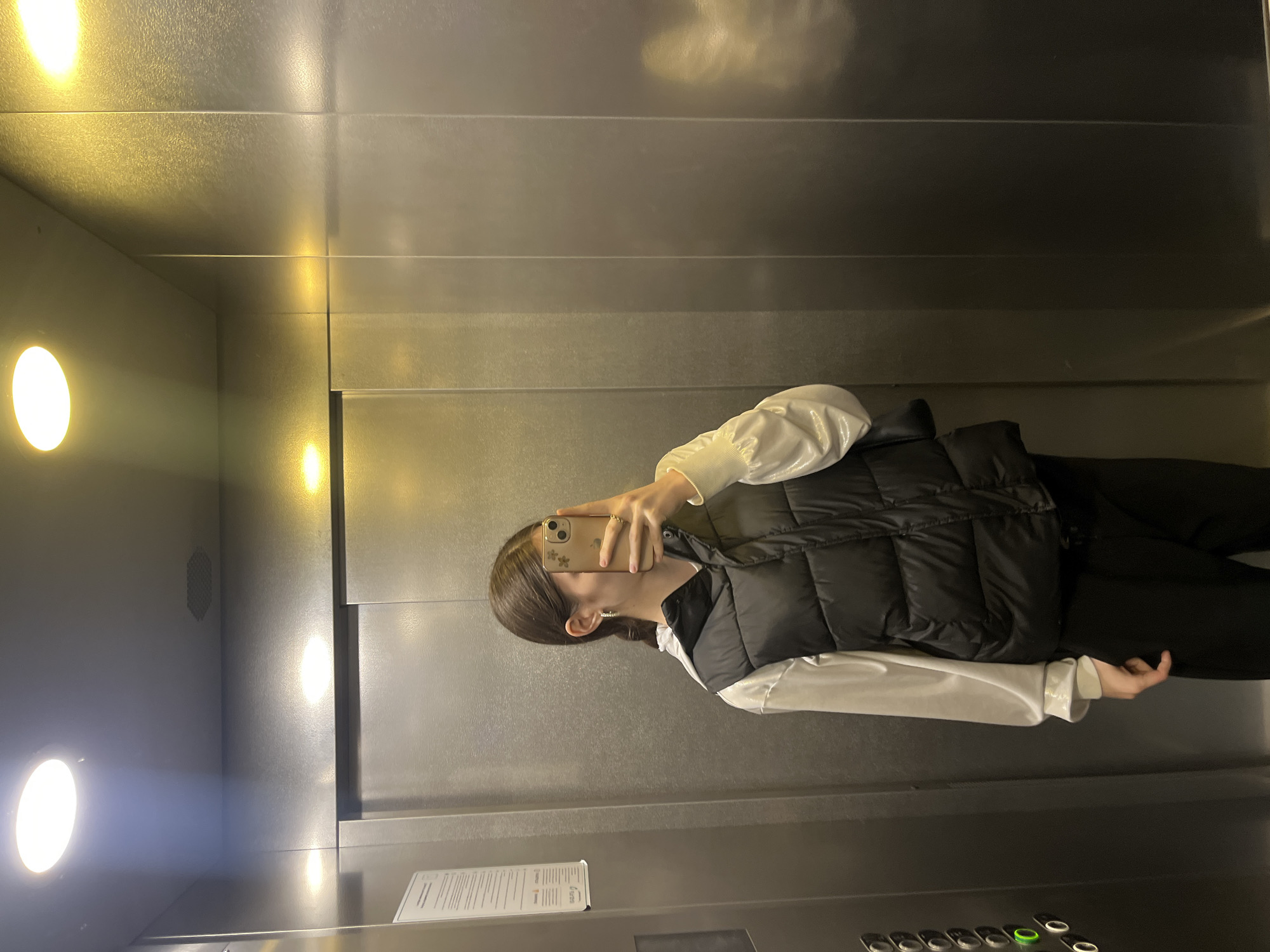 A person with brown hair is taking a photo in an elevator. The elevator has metal walls and ceiling with recessed circular lights. The person is wearing a white long-sleeved shirt and a black quilted vest. The scene is not identifiable as being located in Saint Petersburg, Russia.