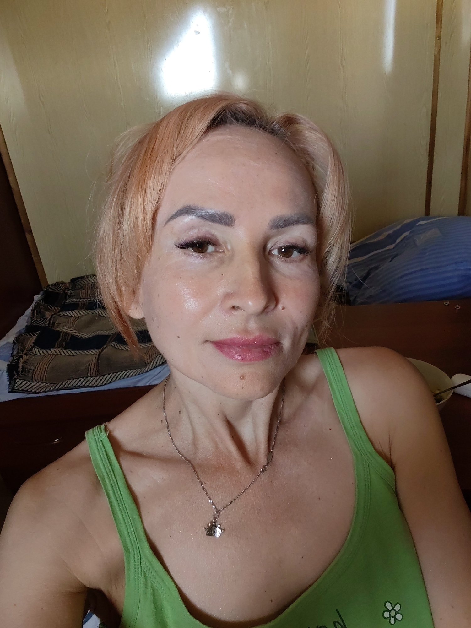 The image shows a close-up of a woman with light brown hair and fair skin. She is wearing a light green tank top and a silver necklace with a pendant. The background appears to be an indoor setting, with a wooden wall and a bed with blue and striped bedding. A patterned blanket is visible on the bed. The lighting suggests an interior space, possibly a bedroom. The image is a selfie, taken from the chest up.