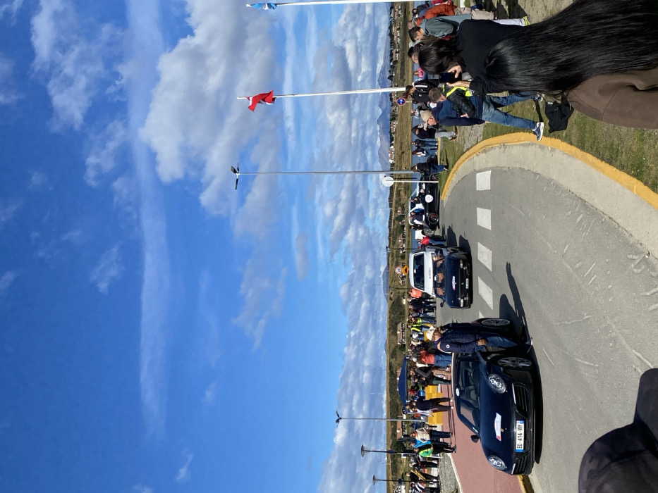 The image depicts a daytime scene in Ushuaia, Argentina, showing a car procession on a paved road adjacent to a body of water, likely the Beagle Channel. The sky is bright blue with scattered white cumulus clouds.
In the foreground, the back of an individual's head, wearing a dark cap, partially obscures the lower left corner. To the right, the back of another person with long dark hair, wearing a brown jacket, is visible.
The main subject is a curved asphalt road with a visible crosswalk in the foreground. On the road, a line of cars is either stationary or moving slowly. The lead vehicle in the immediate foreground is a black Porsche 911 convertible, displaying a white sticker on its front bumper and hood, and an Argentine license plate "ED-414-WY". A man in a white hat and blue jacket stands beside the driver's side of the Porsche. Behind the Porsche is a black Ferrari convertible with its top down, occupied by a driver. Further back in the procession, a white van, a dark-colored pickup truck, and another black car making a right turn are visible. A circular "20" speed limit sign is visible above the white van.
Numerous people are gathered along both sides of the road, observing the vehicles. On the left, a crowd stands on a reddish-paved path. On the right, spectators stand on a grassy verge, some holding mobile phones to photograph the event. The edge of the road on the right is marked with a yellow-painted curb.
In the midground, two tall streetlights with dark fixtures are present. Further to the right, two tall flagpoles display flags; one is predominantly red, and the other features light blue and white stripes (consistent with the flag of Tierra del Fuego province).
The background reveals a body of water with buildings lining the distant shore, characterized by light-colored walls and dark or red roofs. Beyond the settlement, a range of mountains is visible under the partially cloudy sky.