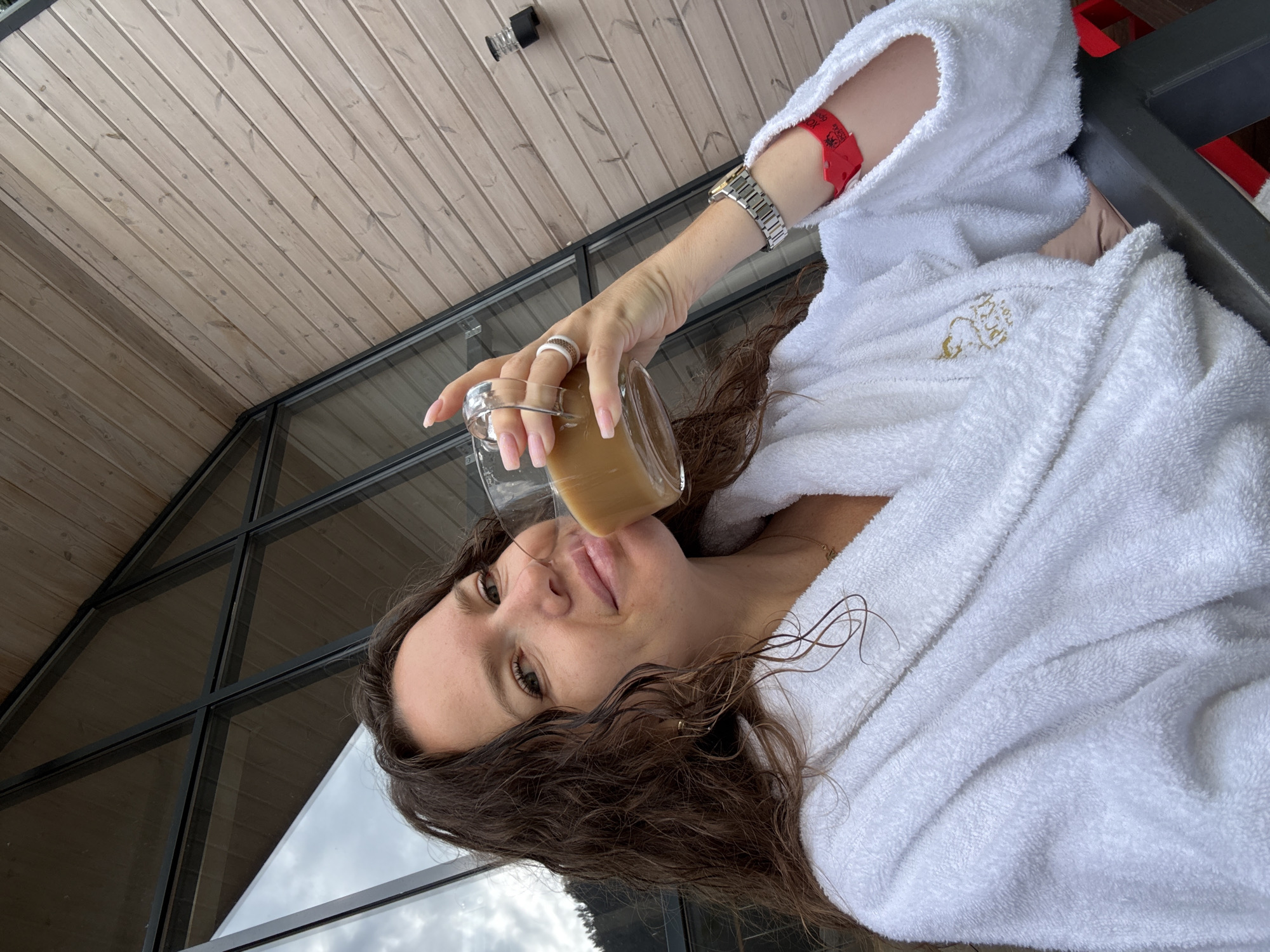 A woman with curly brown hair, wearing a white bathrobe, holds a glass mug filled with coffee. She is outdoors, with a wooden-paneled wall and a large window with a dark frame in the background. The window reflects a cloudy sky and a treed hillside. On the woman's left hand, she wears two rings, and on her left wrist, she wears a watch. A red wristband is visible on her right forearm. A golden embroidered logo is visible on the left side of the bathrobe. The scene appears to be in Barangol, Russia.